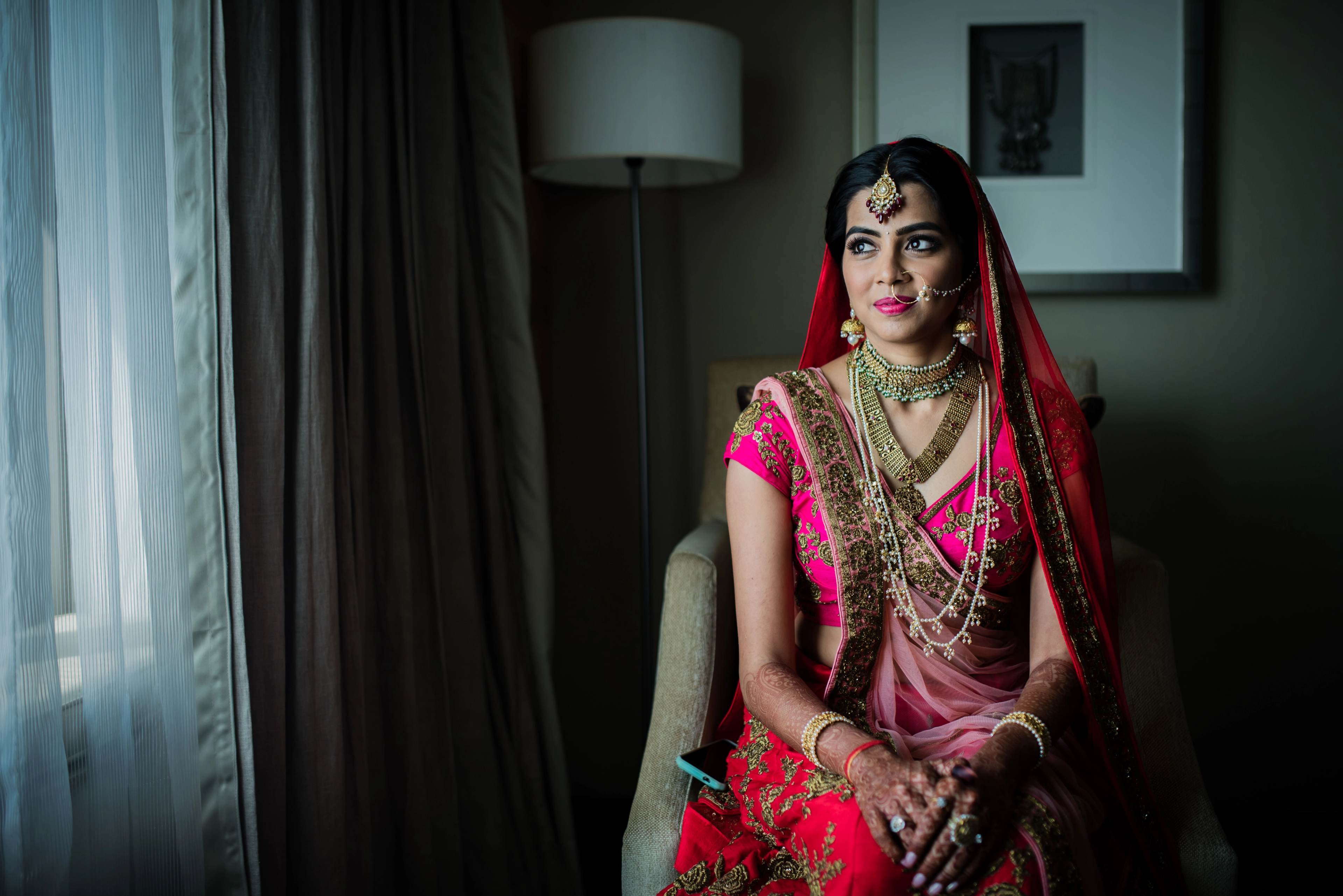 Woman in pink and red traditional bridal attire with veil, jewelry, and henna, seated indoors