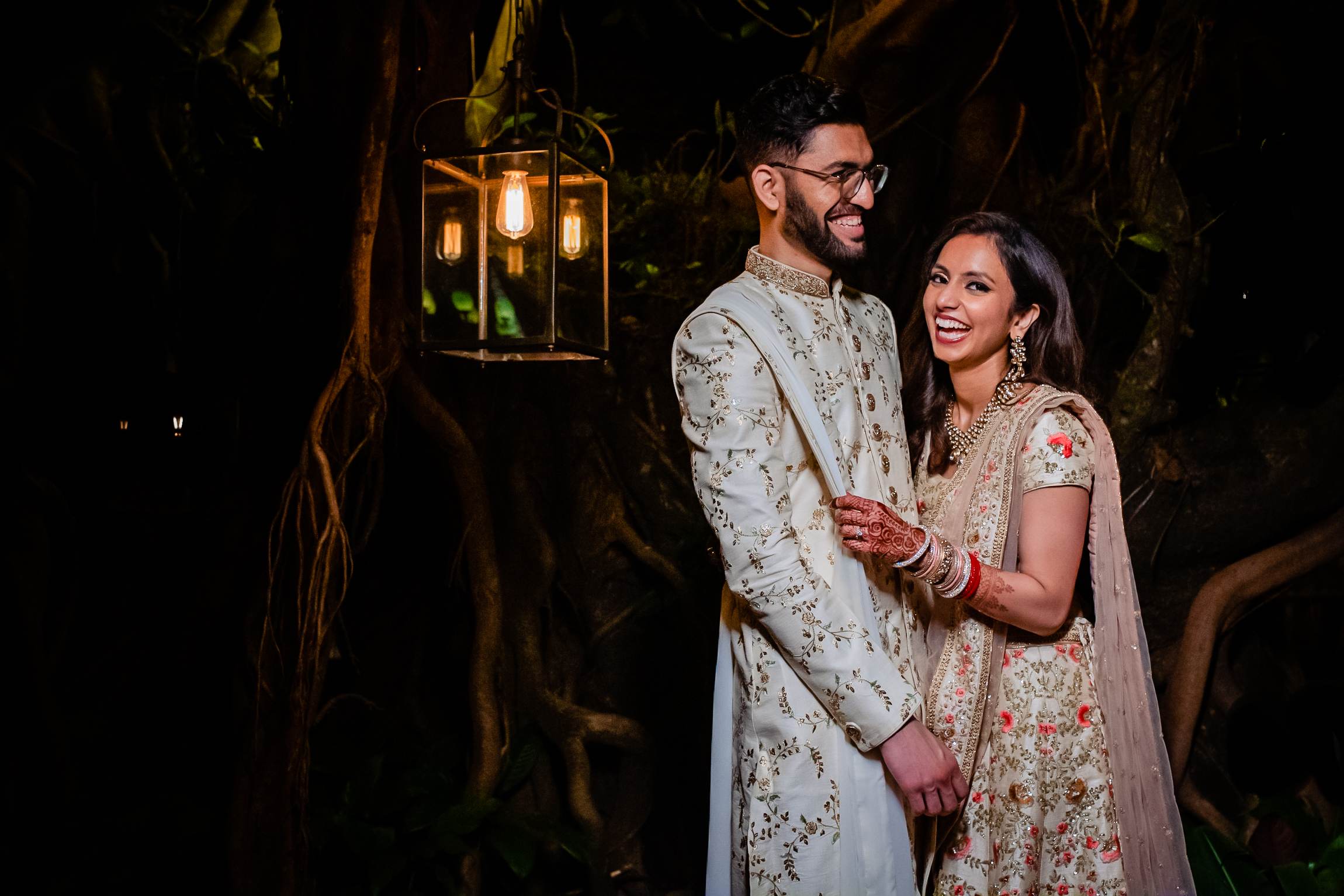Avani and Srikant stand together in ornate traditional attire under glowing lanterns in Goa's nighttime setting