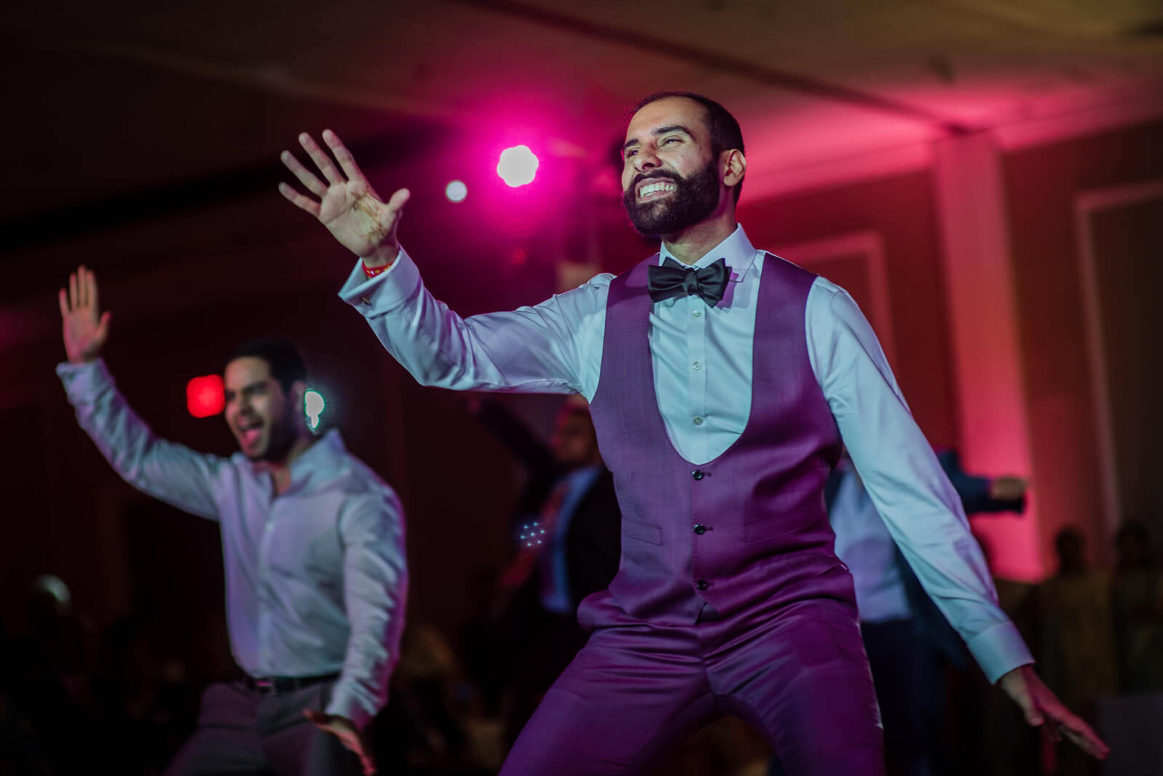 Ankit waves to guests while dancing in purple vest at New York reception with pink uplighting