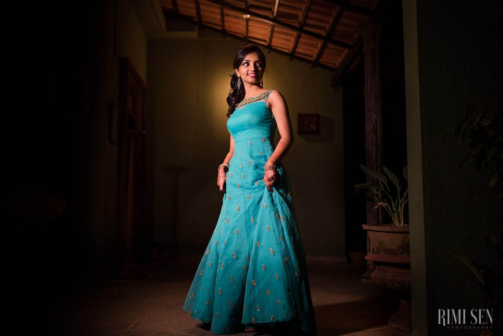 Pooja stands in a turquoise embroidered gown in an indoor colonial setting, warm light illuminating her elegant pose