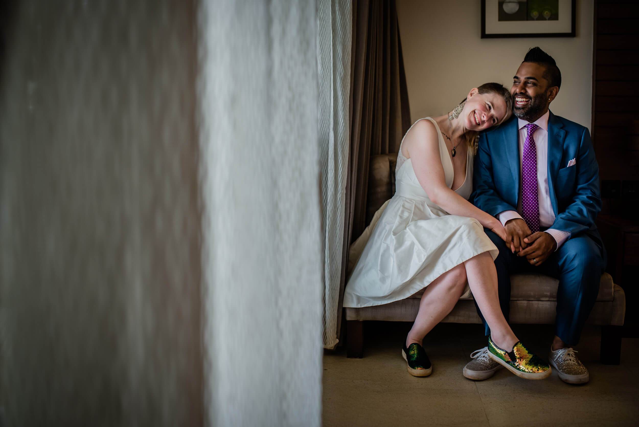 Sarah and Ravin sit together laughing in a hotel room, holding hands in casual wedding attire