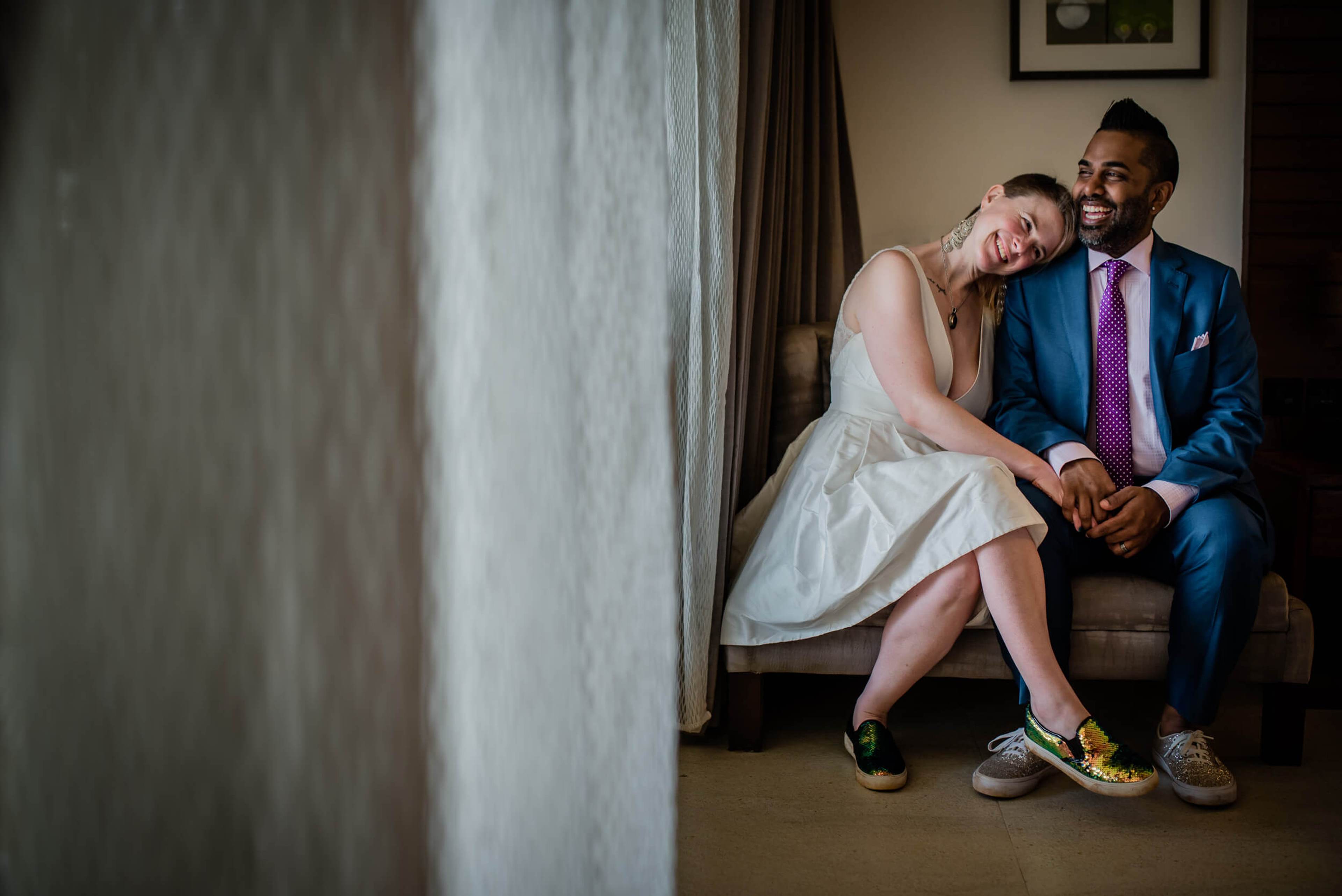 Sarah and Ravin sit together laughing in a hotel room, holding hands in casual wedding attire