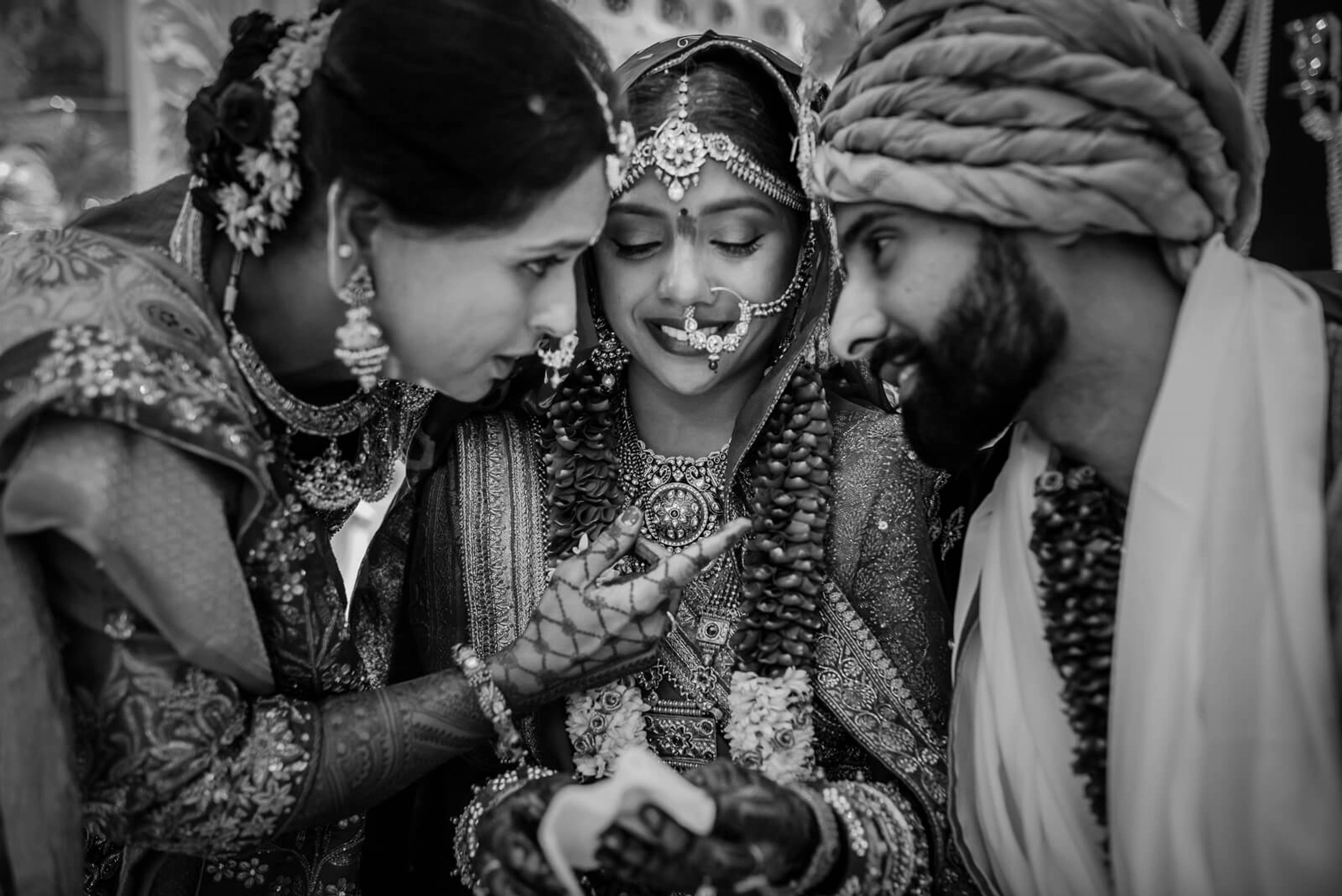Bride in ornate lehenga surrounded by joyful family members in traditional Indian wedding attire, black and white portrait