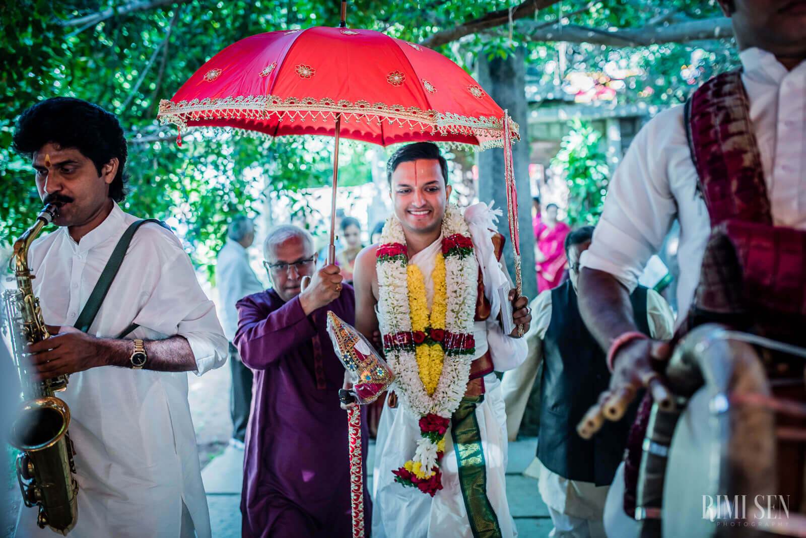 Groom Aniket under red umbrella with flower garland, saxophonist and family at Bangalore wedding
