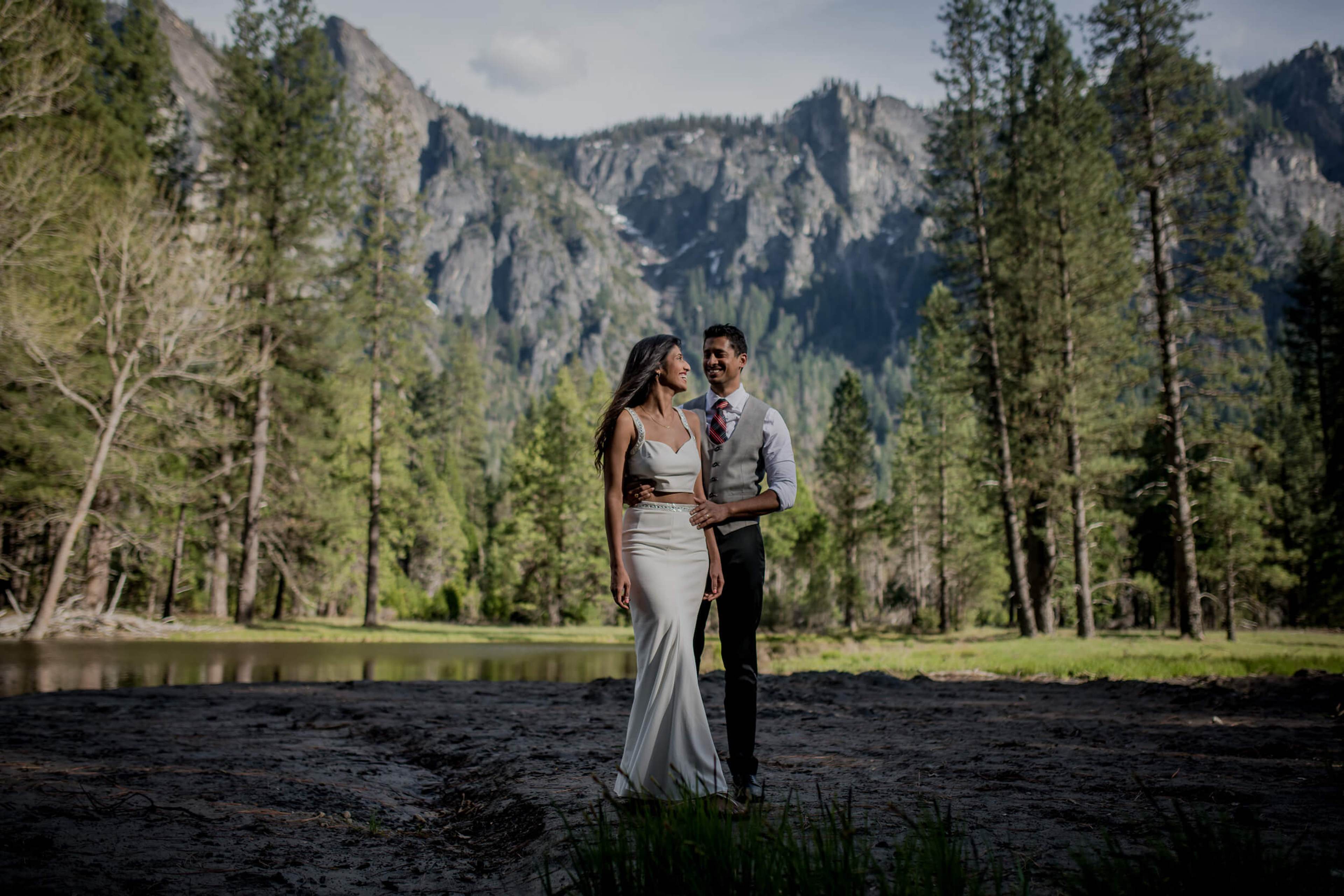 Anu and Raghu embrace by a river, forested mountains and evergreens behind them in California