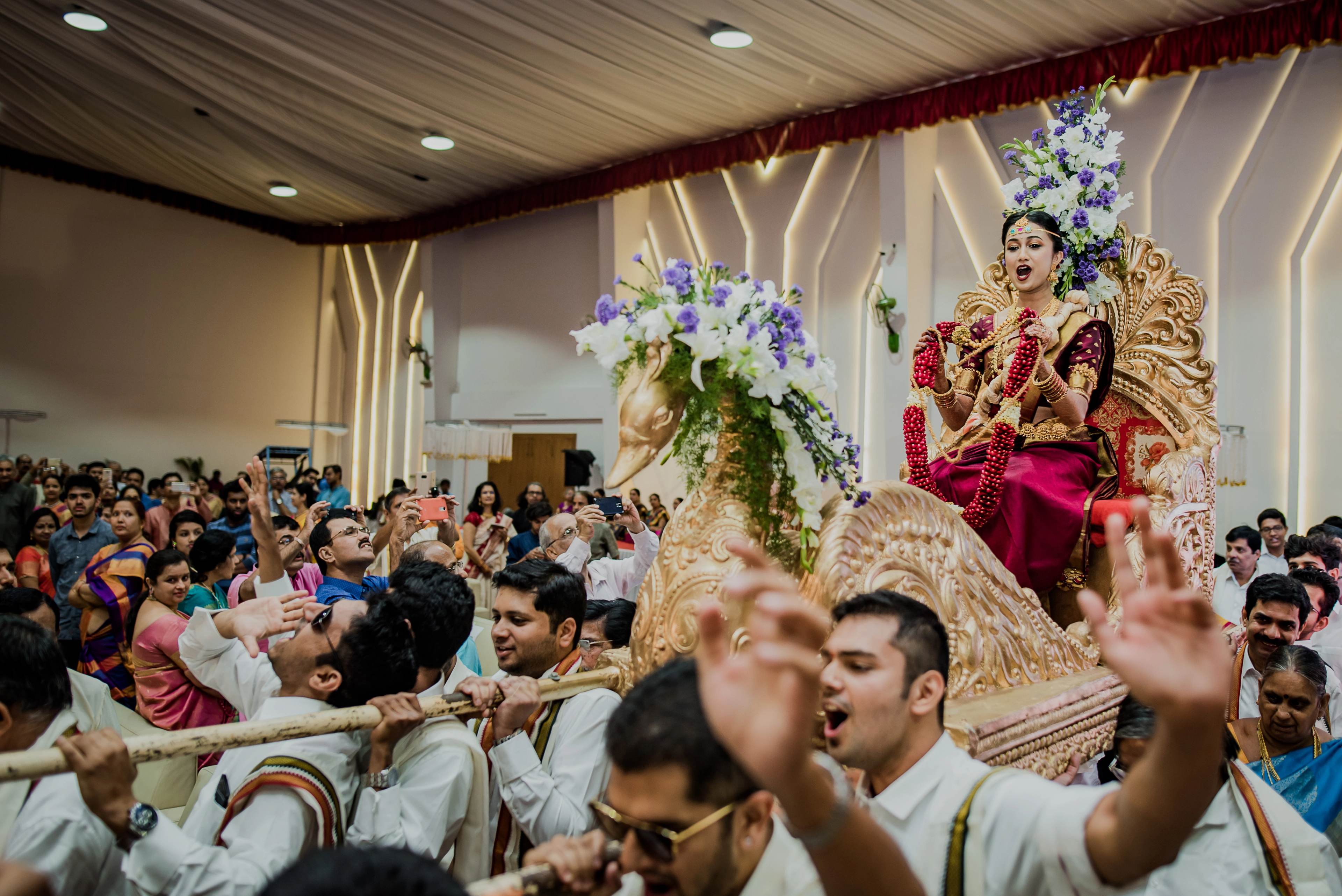 Bride in red and gold lehenga seated on ornate swing above celebrating guests in traditional attire indoors