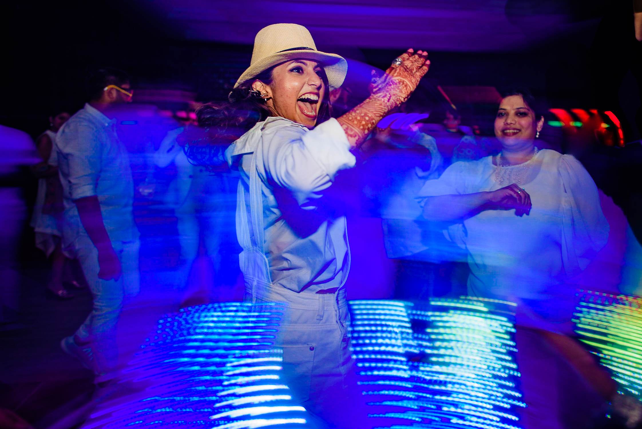 Avani dancing in white suit and hat under blue stage lights at Goa wedding reception, guests celebrating nearby