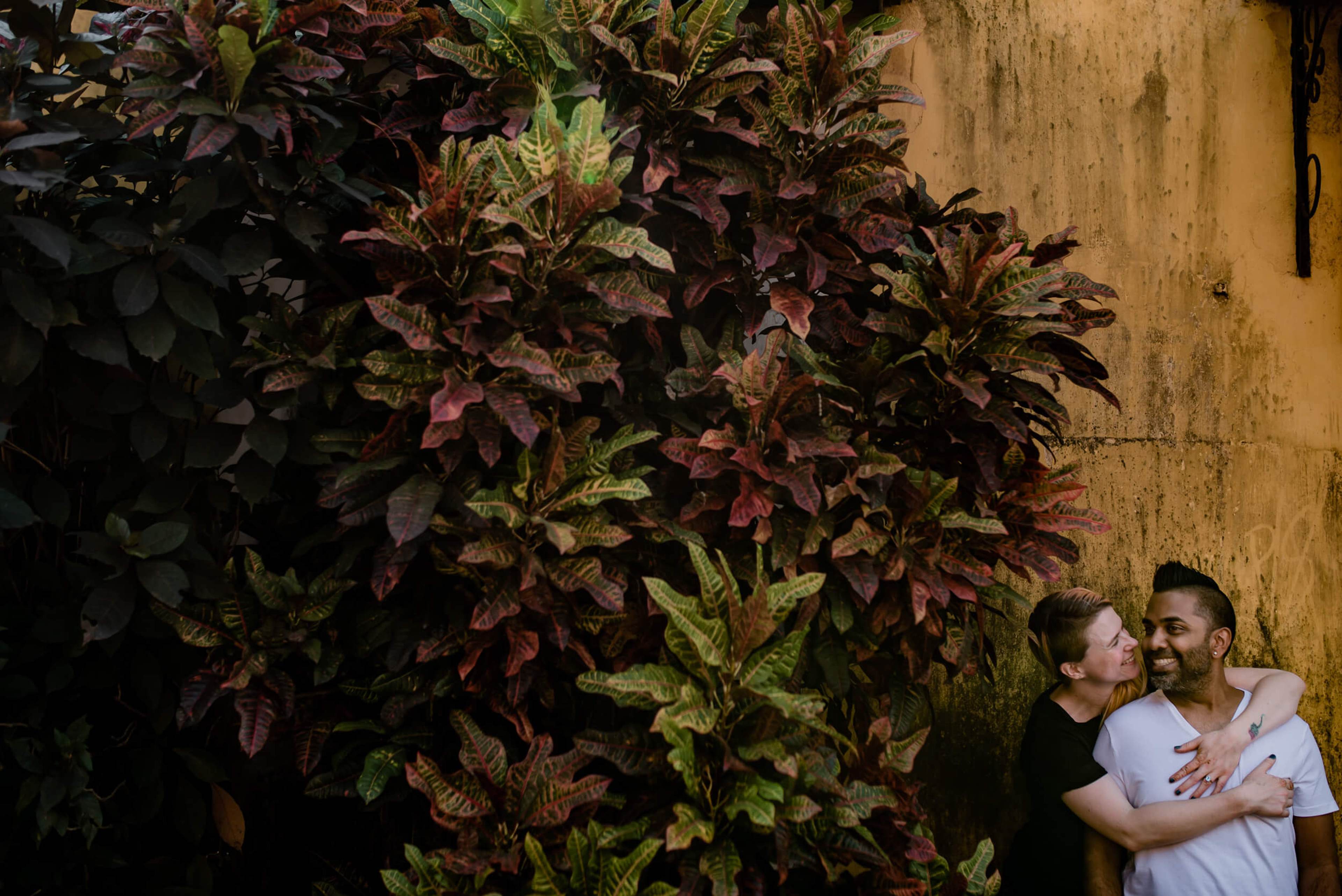 Sarah and Ravin embrace by lush climbing vines against weathered yellow wall in Goa