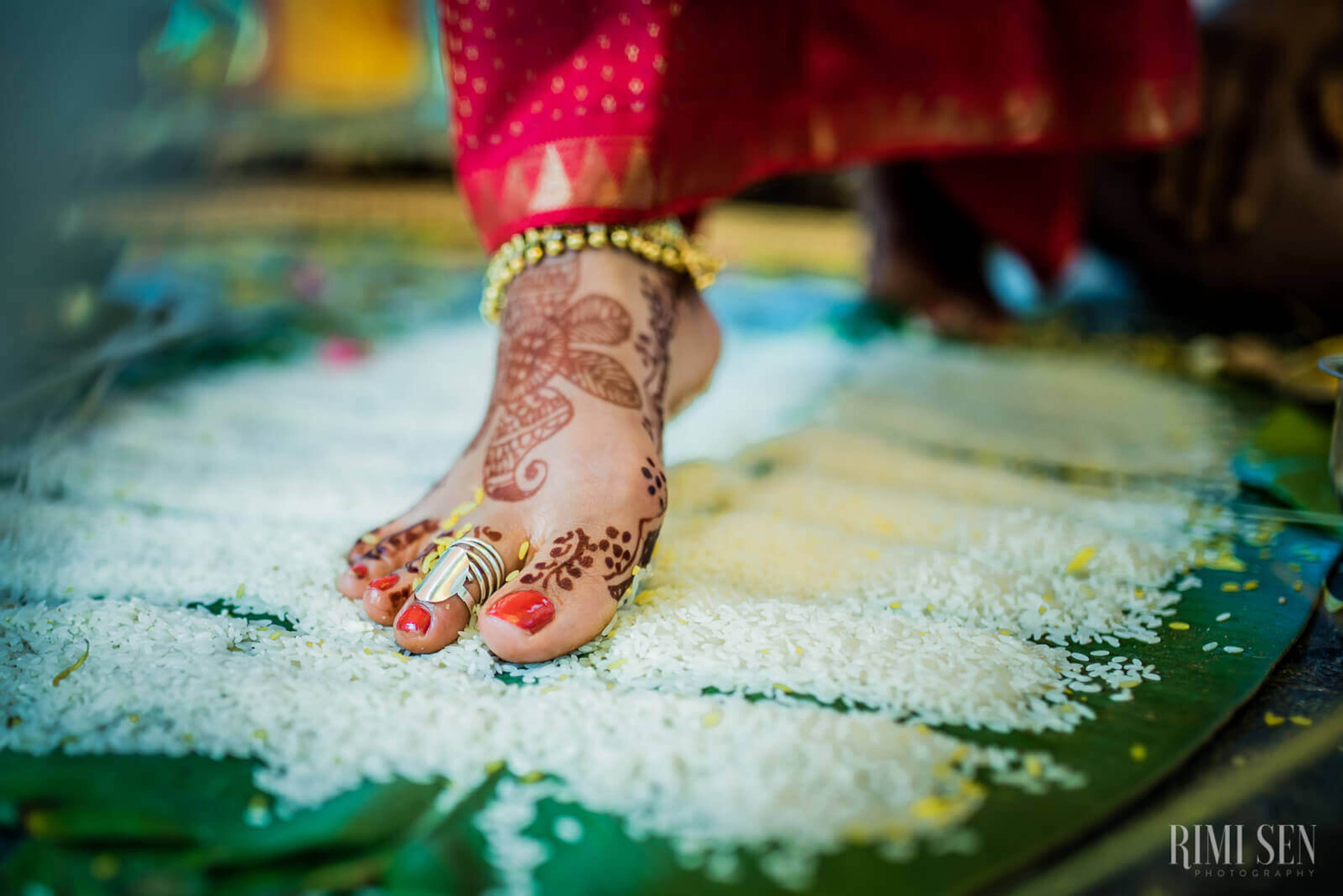 Pooja's henna-decorated foot with gold anklet and red nail polish on colorful floor detail