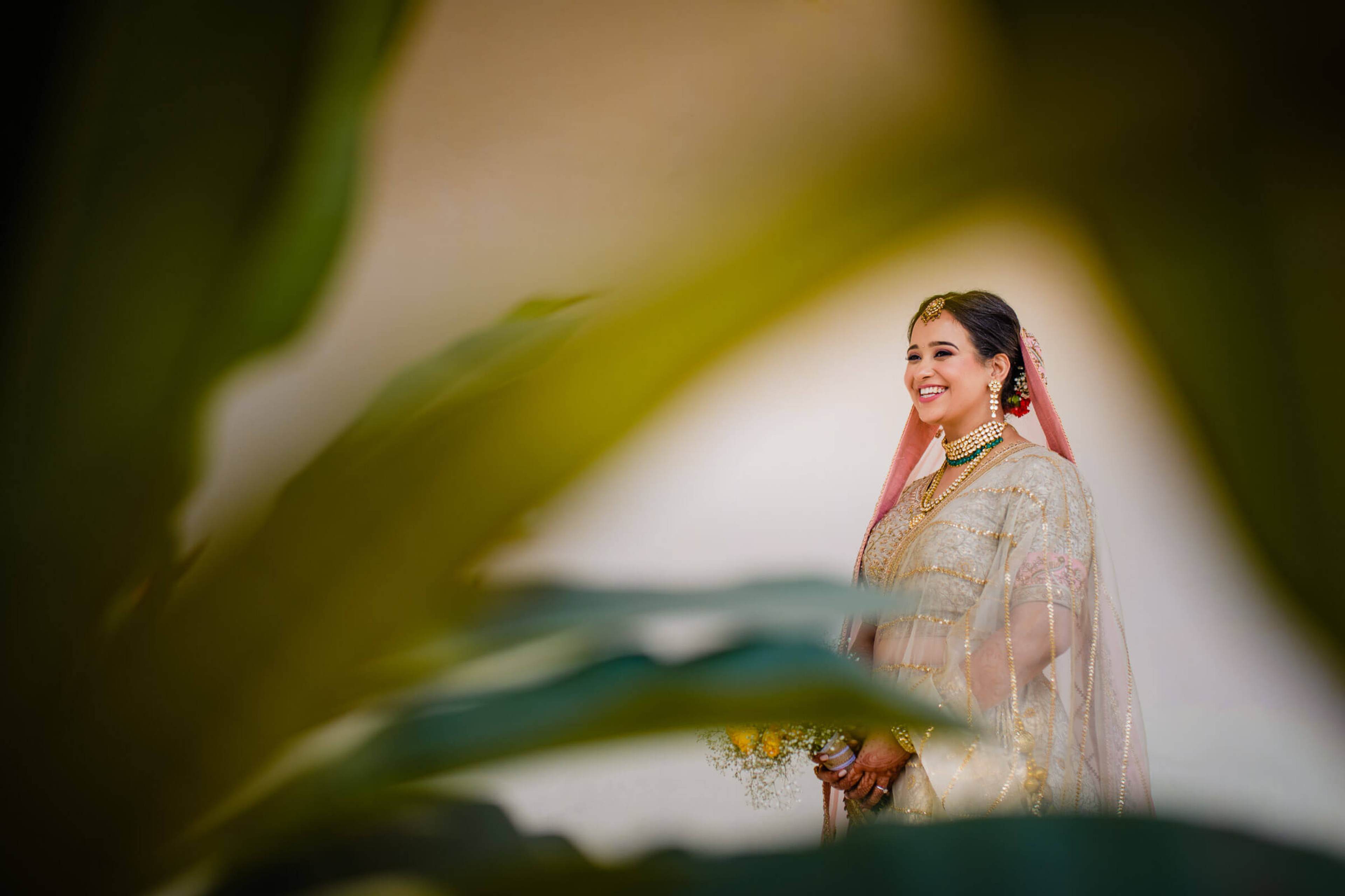 Maitrayee in gold embroidered lehenga and pink veil, framed by green foliage in Rajasthan