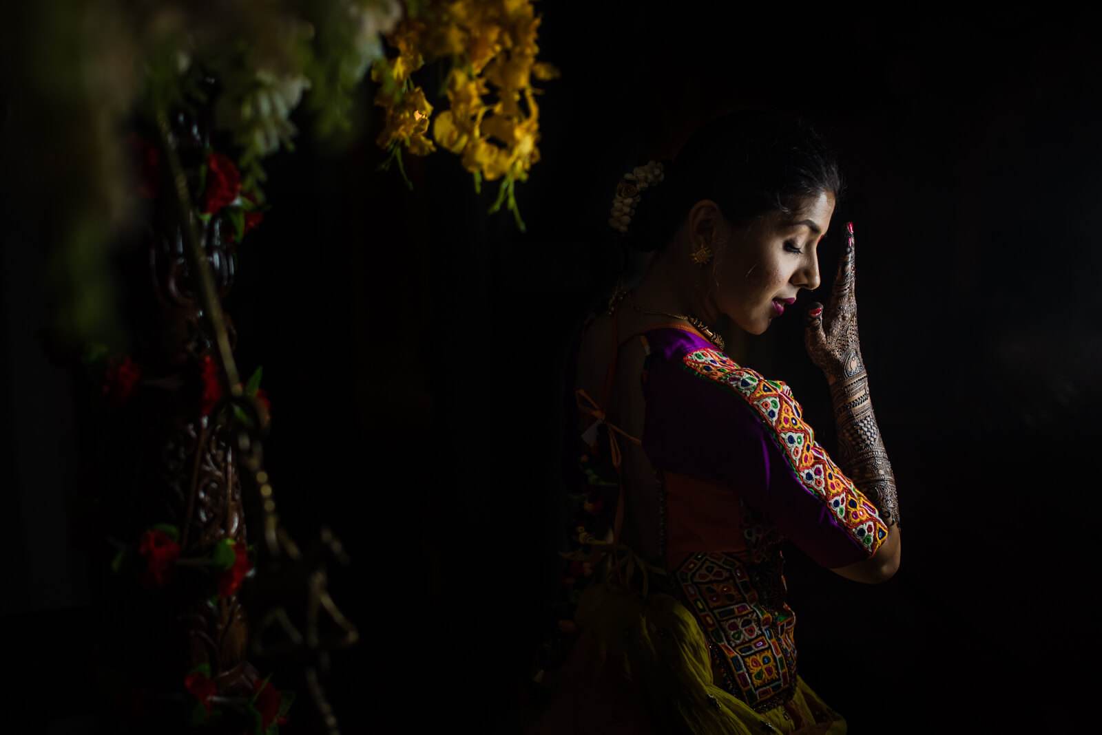 Barkha in embroidered burgundy attire, dramatic side-lit portrait against dark background with hanging flowers