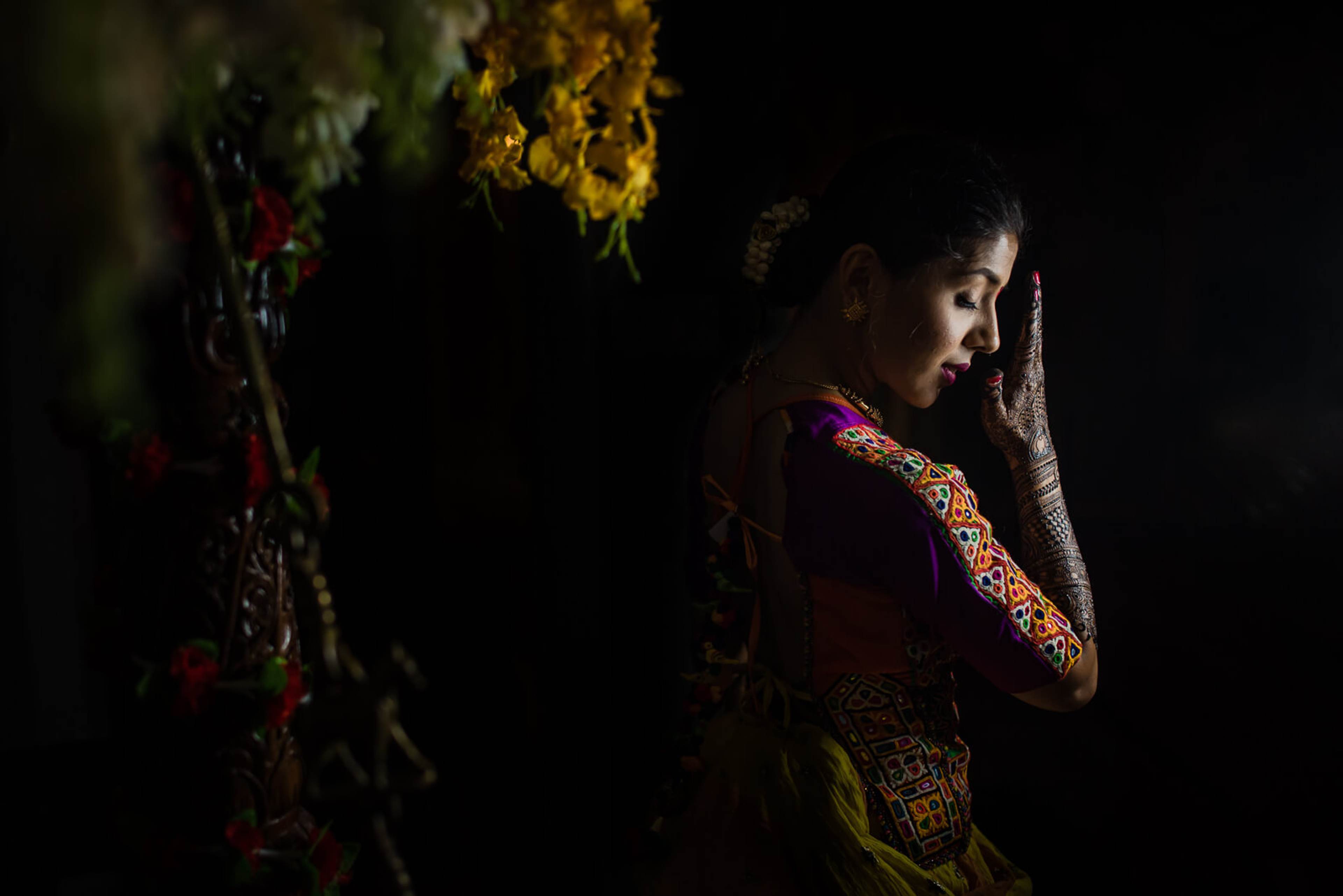 Barkha in embroidered burgundy attire, dramatic side-lit portrait against dark background with hanging flowers