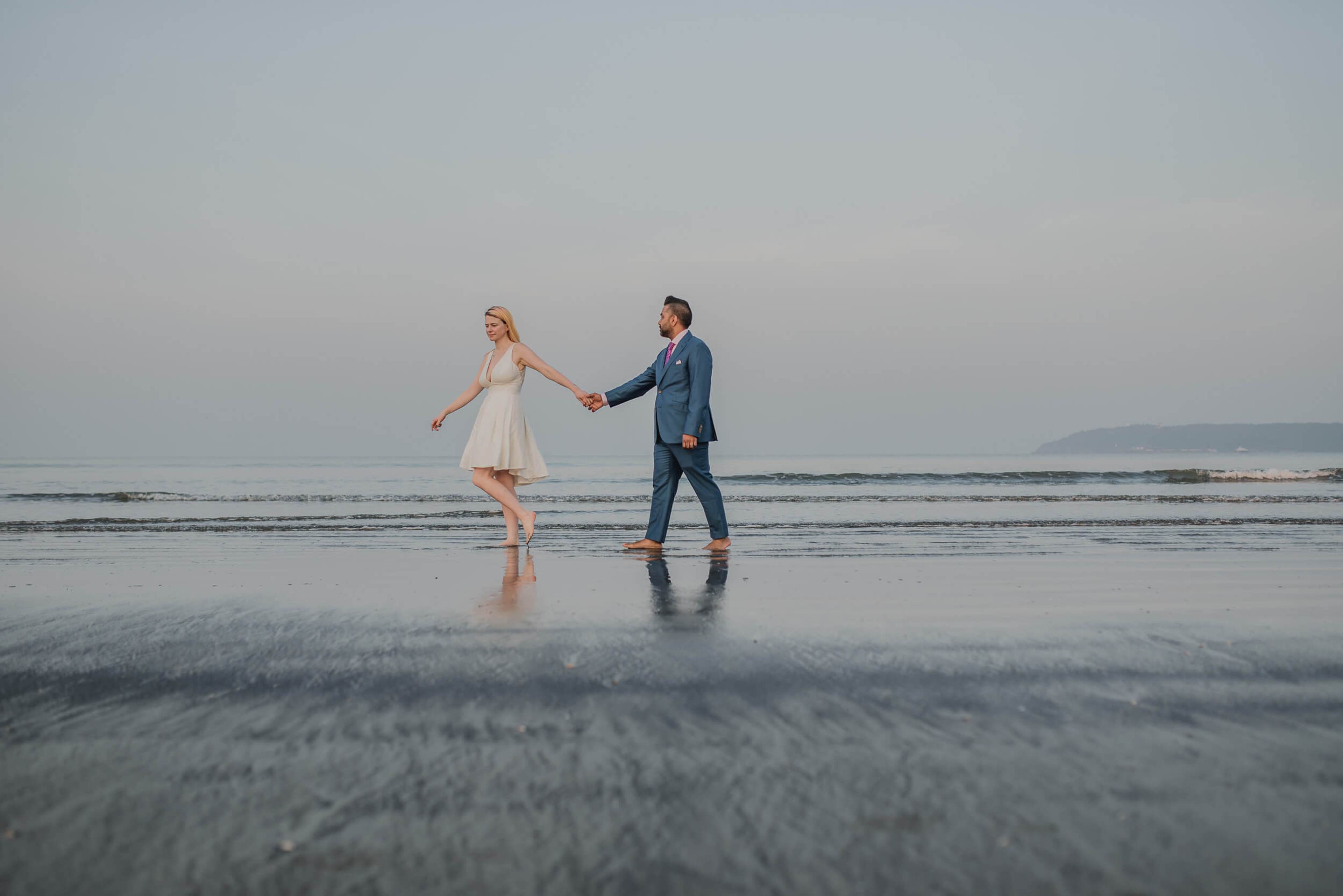 Sarah and Ravin hold hands wading in shallow ocean water at Goa beach at dusk