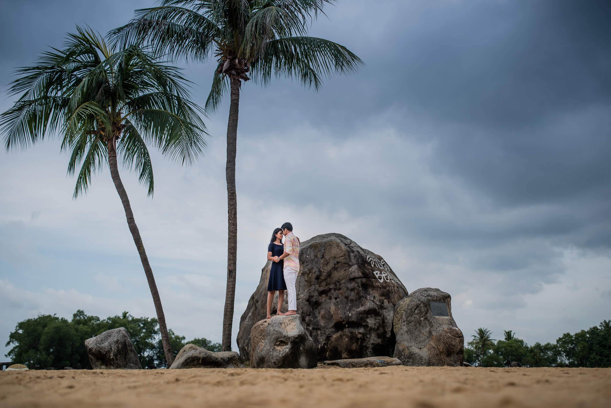 Yash and Disha embrace on a granite boulder beneath tall palms on a Singapore beach
