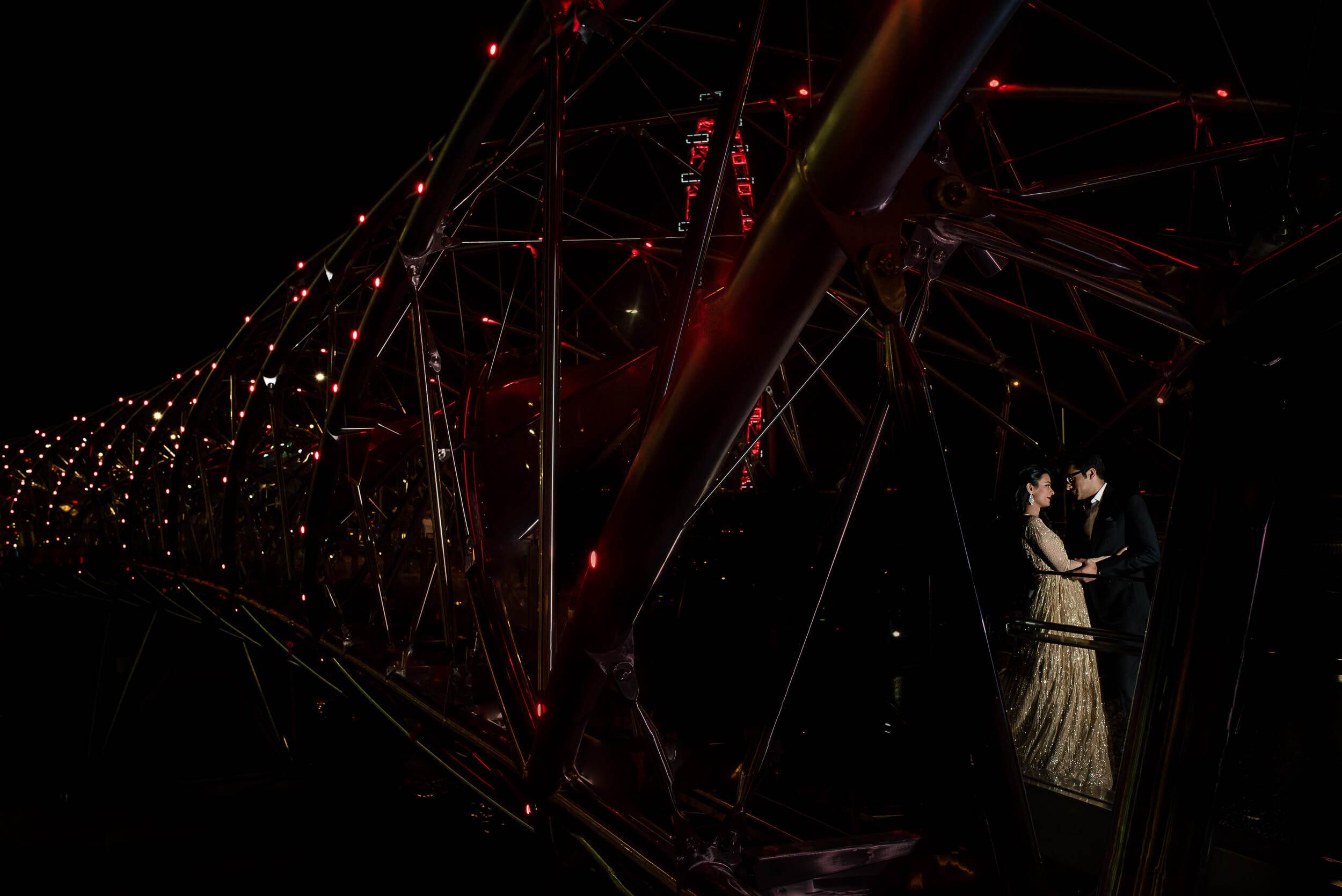 Yash and Disha embrace beneath red-lit industrial structure, intimate moment against Singapore's night skyline