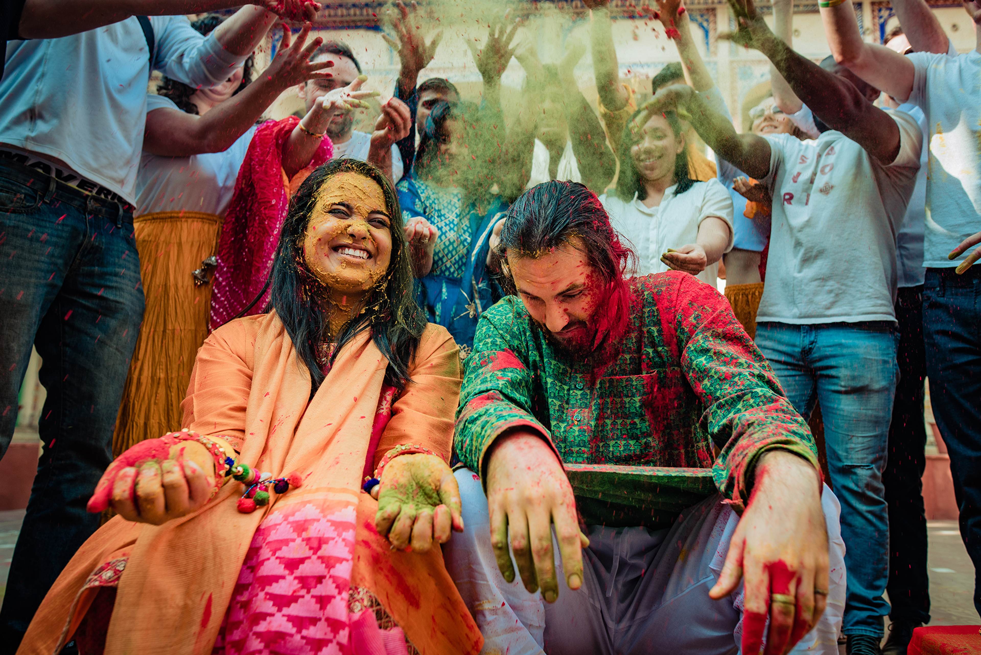 Couple covered in colorful powder celebrating Holi with smiling guests surrounding them in festive attire