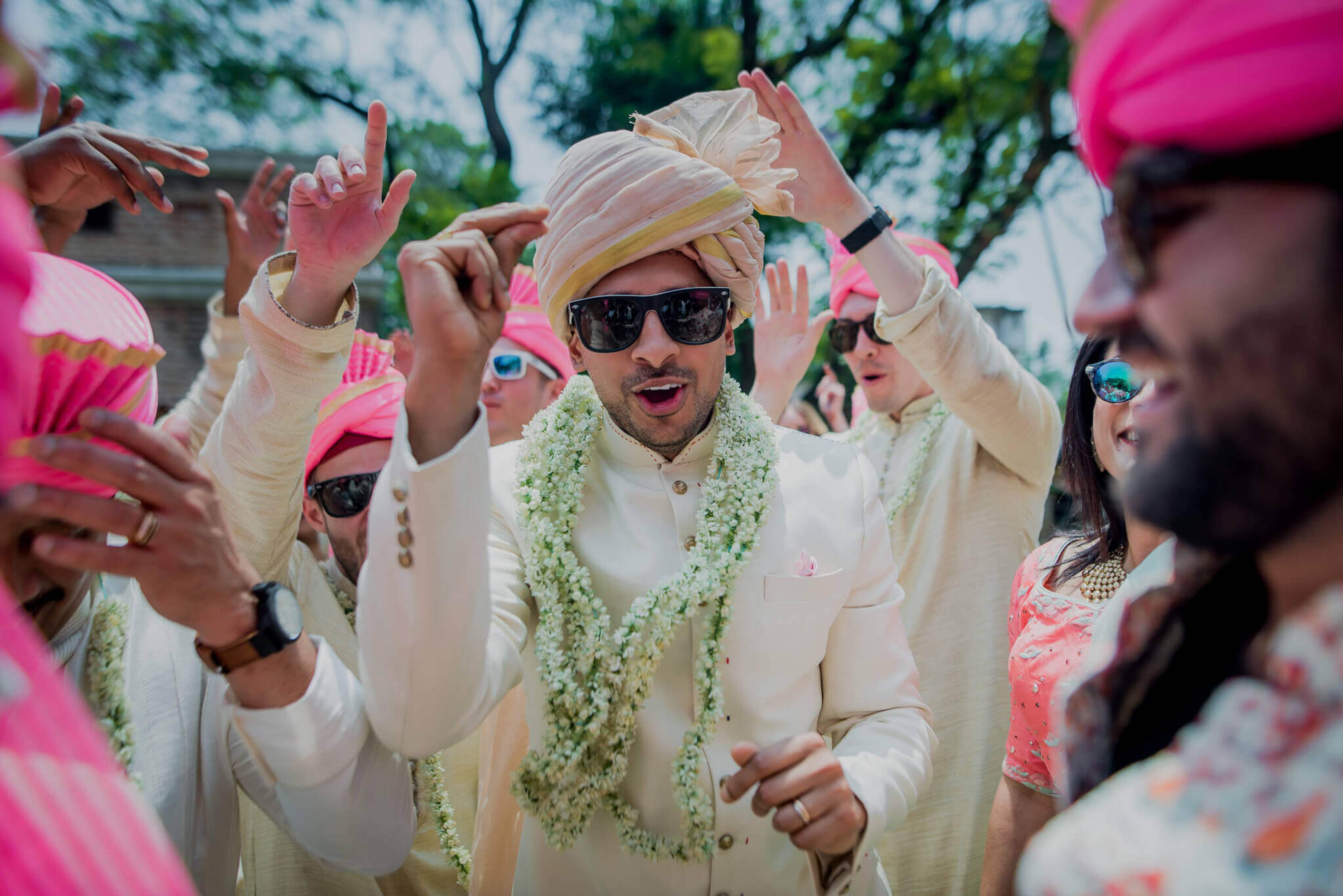 Groom in cream sherwani, sunglasses, and floral garland celebrates with guests in pink and white at Nepal wedding