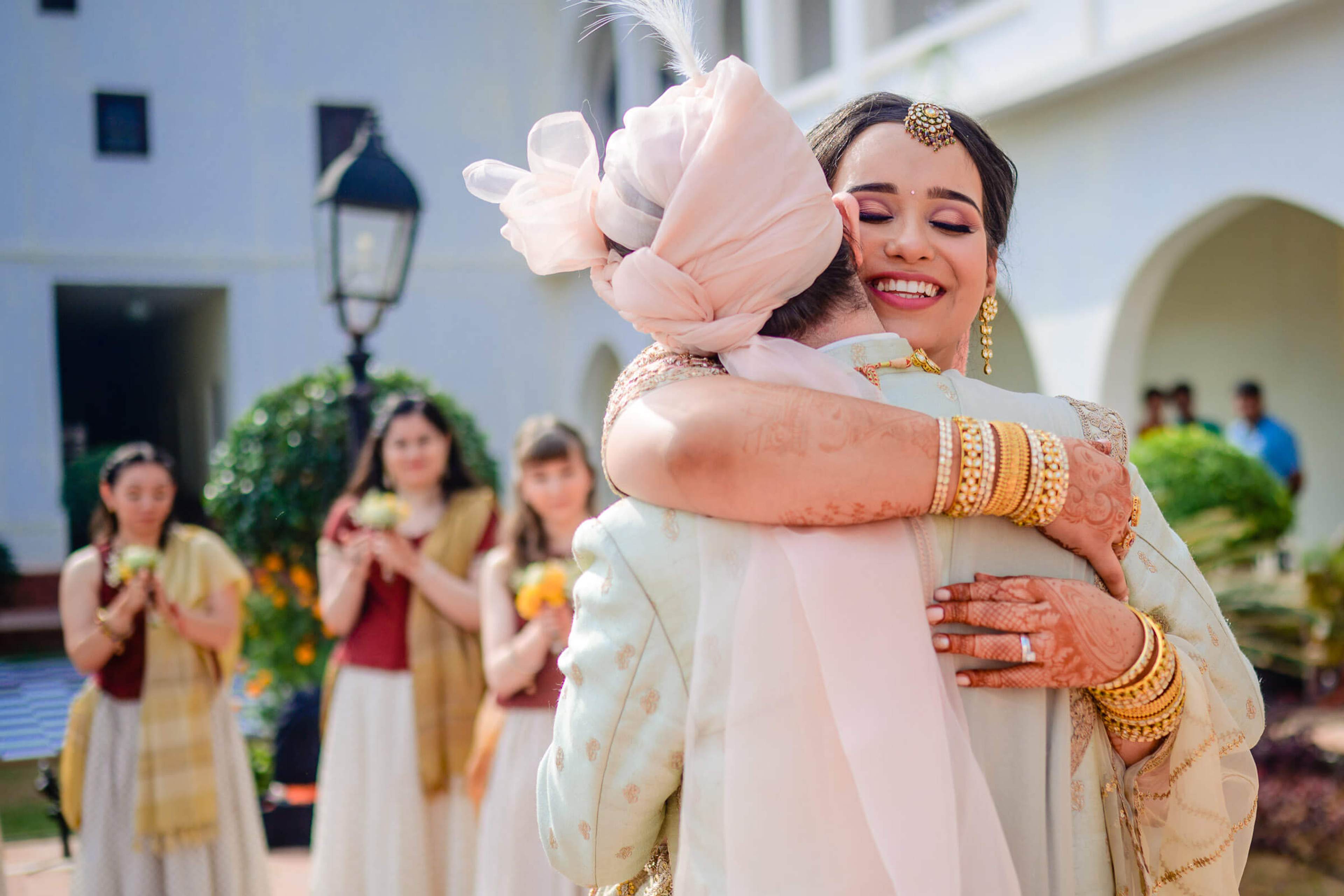 Maitrayee embraces a guest in cream and gold attire, henna on her arms, courtyard celebration in Rajasthan
