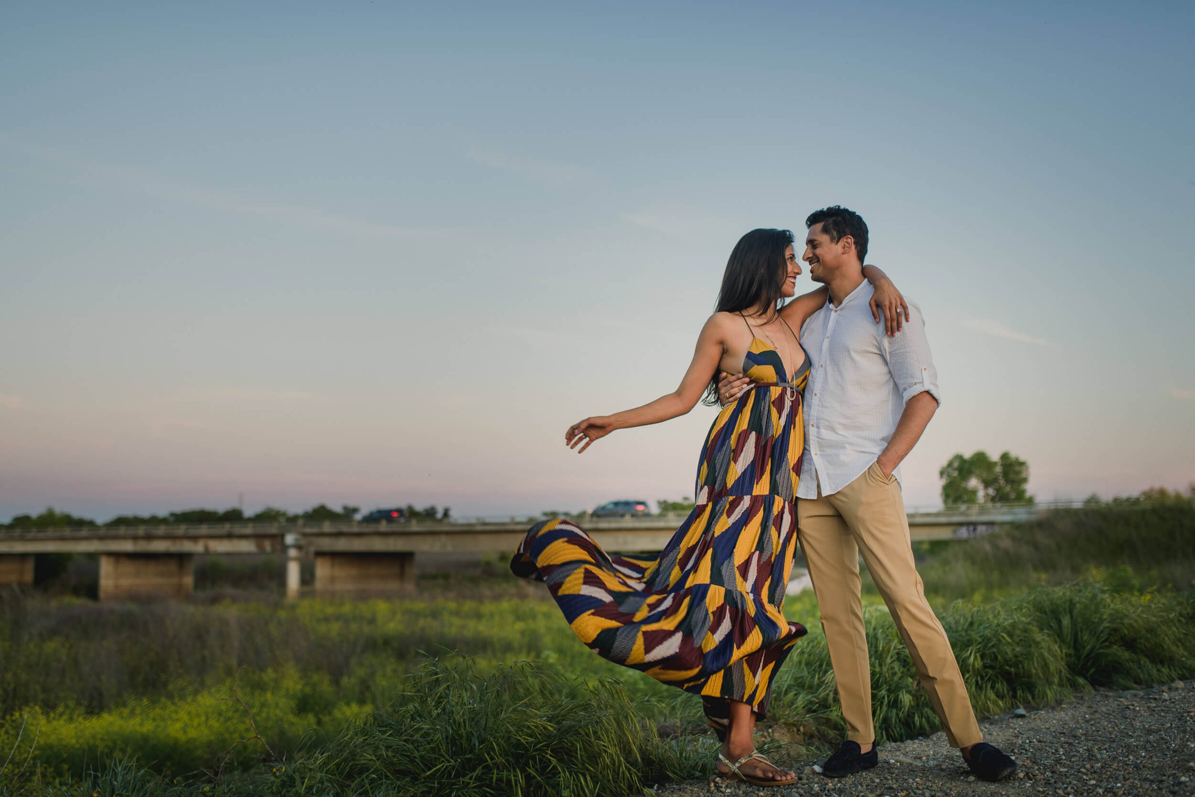 Anu and Raghu embrace on a rural road at dusk, her patterned dress flowing in the wind, California farmland behind them