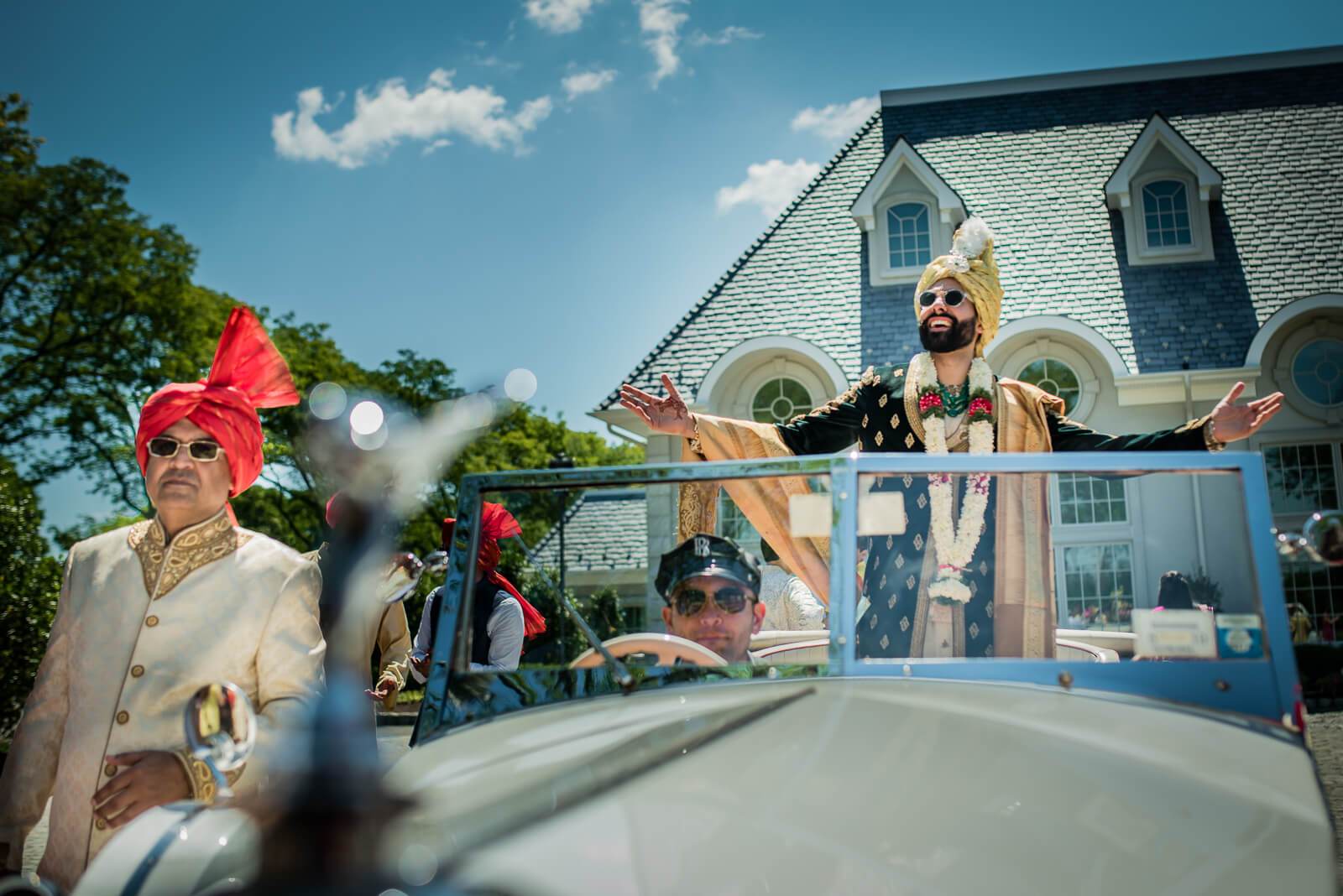 Barkha and Ankit in vibrant traditional attire waving from convertible, New York mansion backdrop