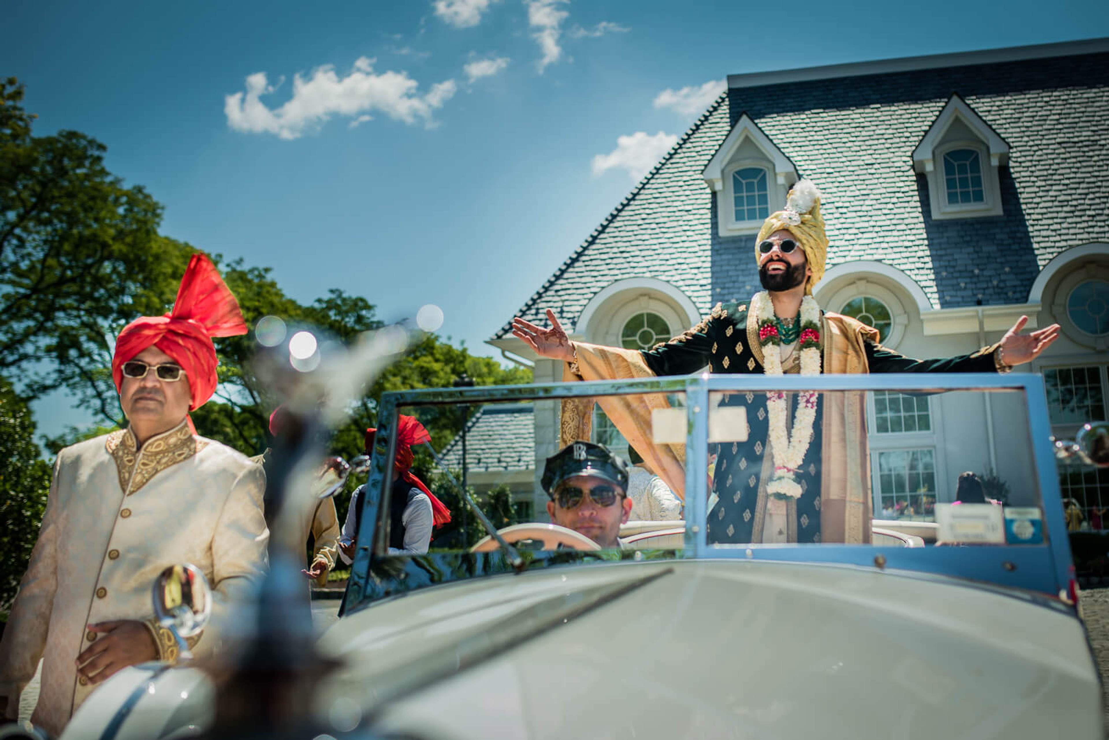 Barkha and Ankit in vibrant traditional attire waving from convertible, New York mansion backdrop