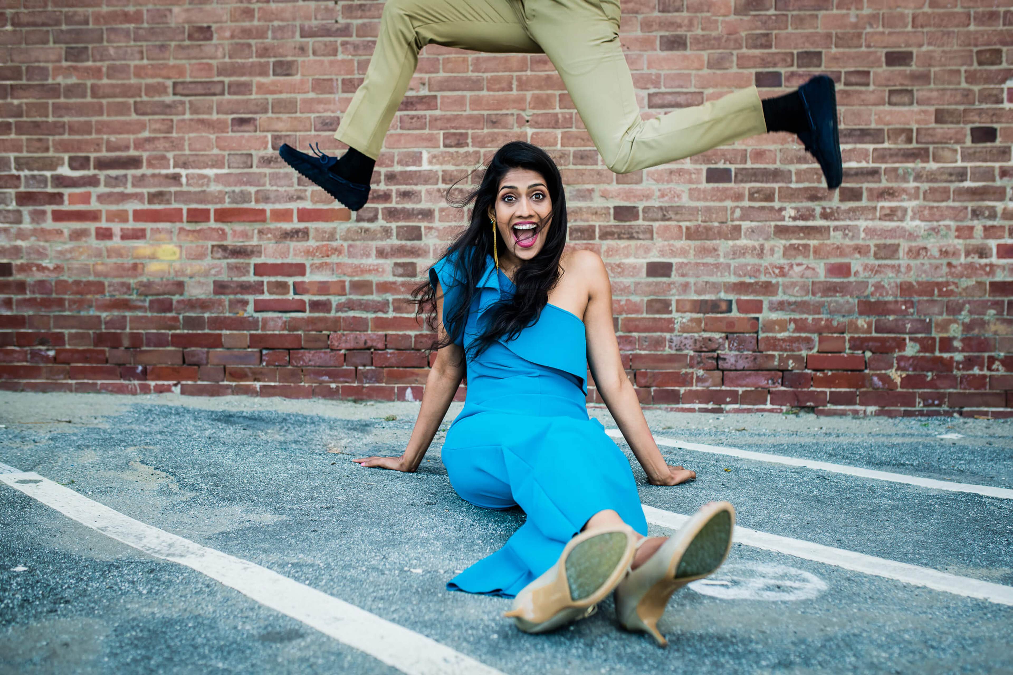 Anu laughs joyfully while sitting in a parking space, legs kicked up, wearing a bright blue dress against a red brick wall