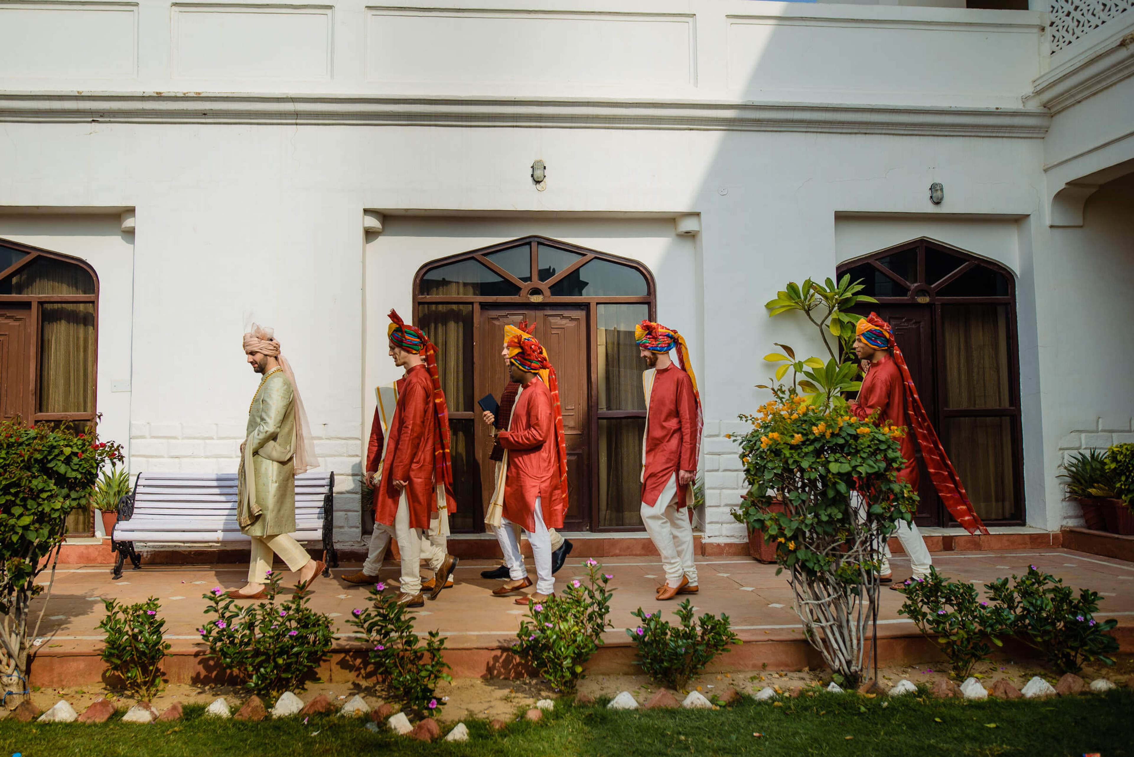 Maitrayee and Eric in traditional red and gold attire walking across a colonial-style Rajasthan courtyard with white walls and potted plants