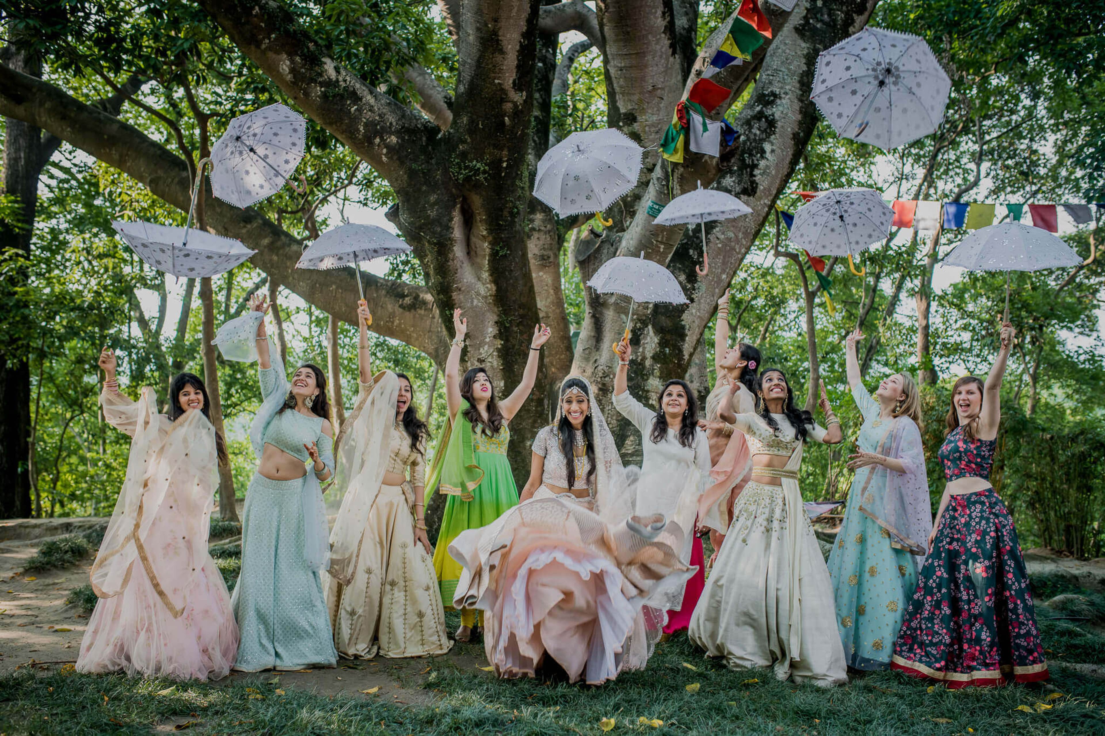 Large group of women in colorful dresses holding umbrellas beneath a sprawling tree with paper lanterns