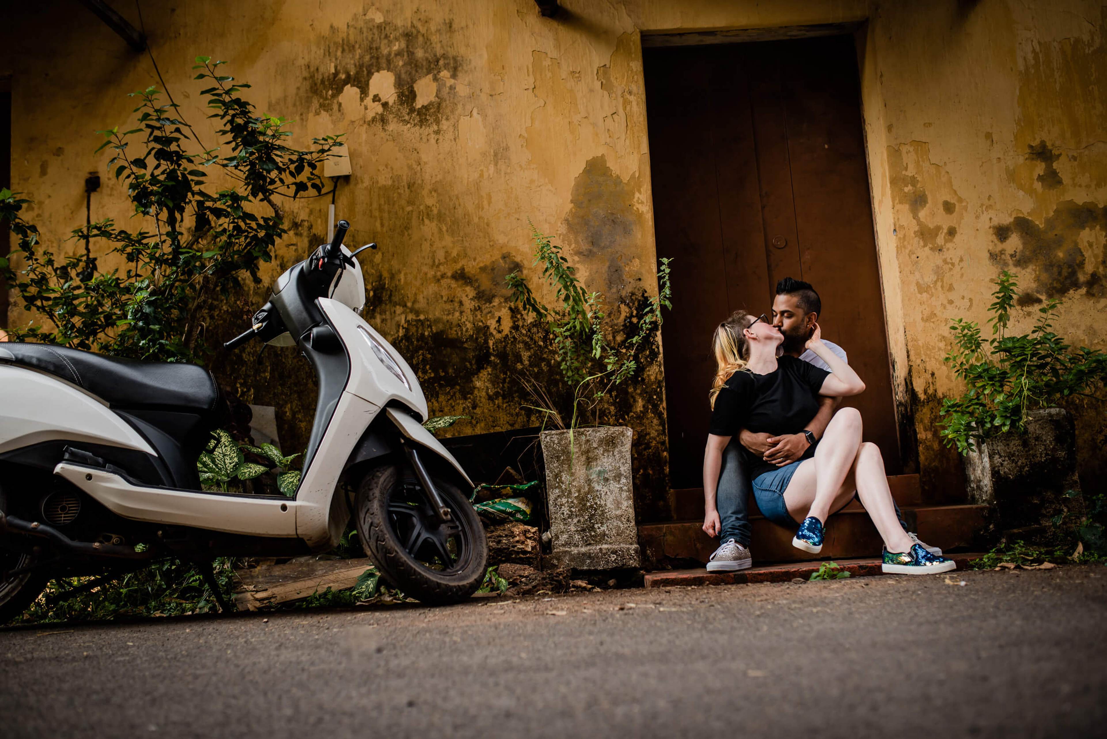 Sarah and Ravin sit by a weathered yellow wall with a white scooter in Goa, warm golden light