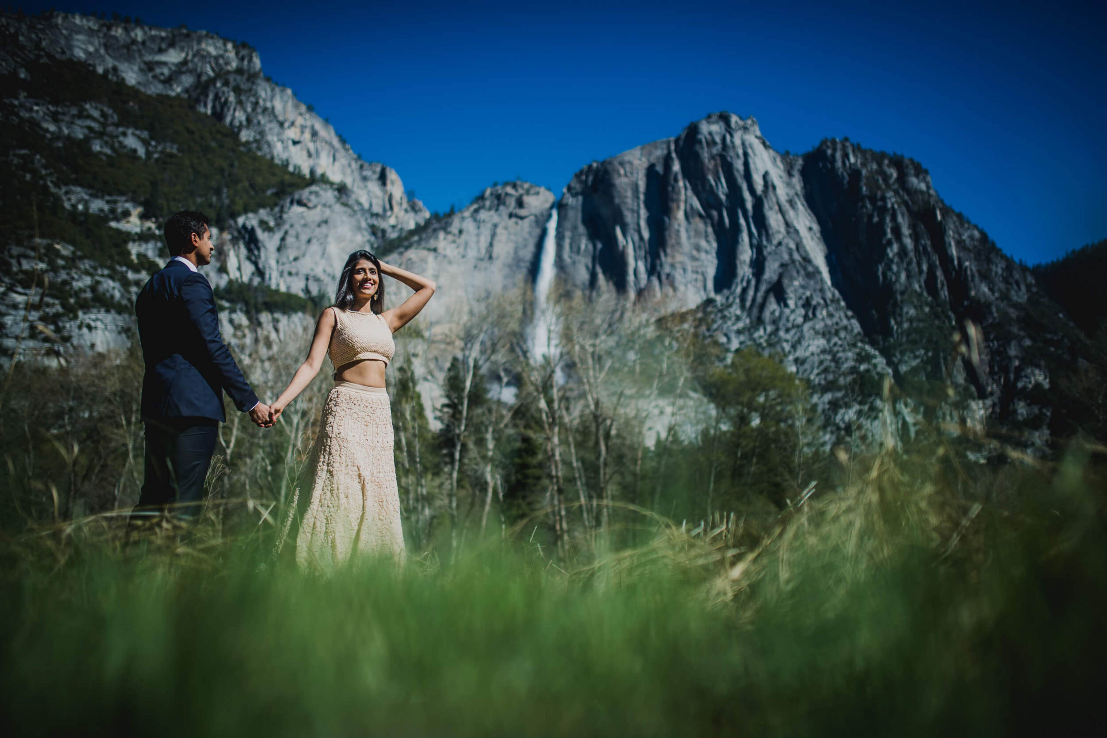 Anu and Raghu hold hands in meadow grass with Yosemite granite cliffs and waterfall behind, clear blue sky