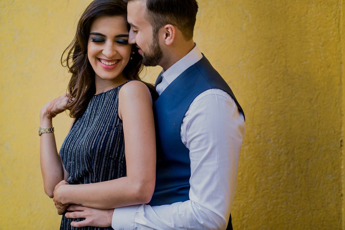 Shreya and Yash embrace against a golden yellow wall, smiling warmly in formal attire