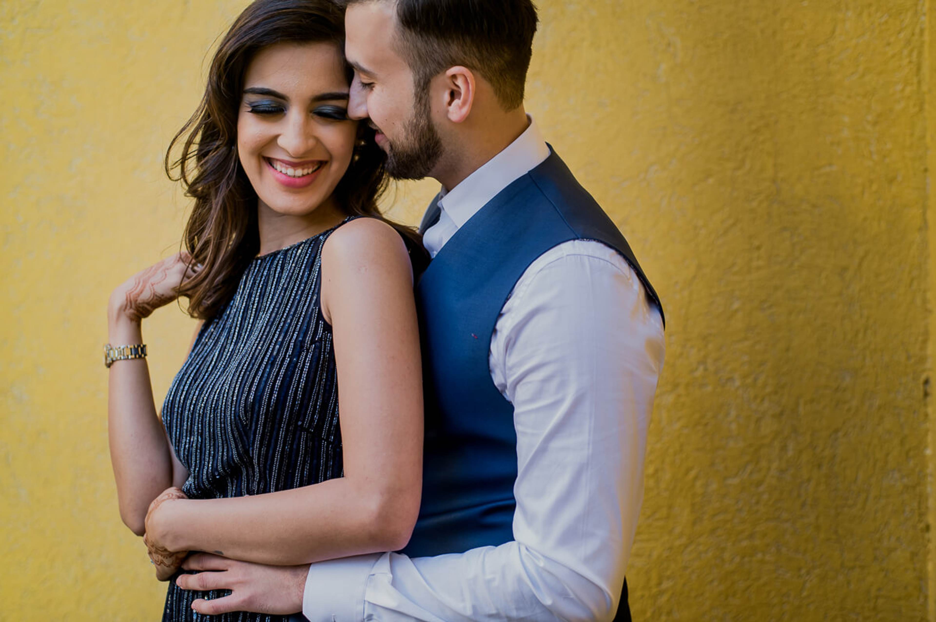 Shreya and Yash embrace against a golden yellow wall, smiling warmly in formal attire