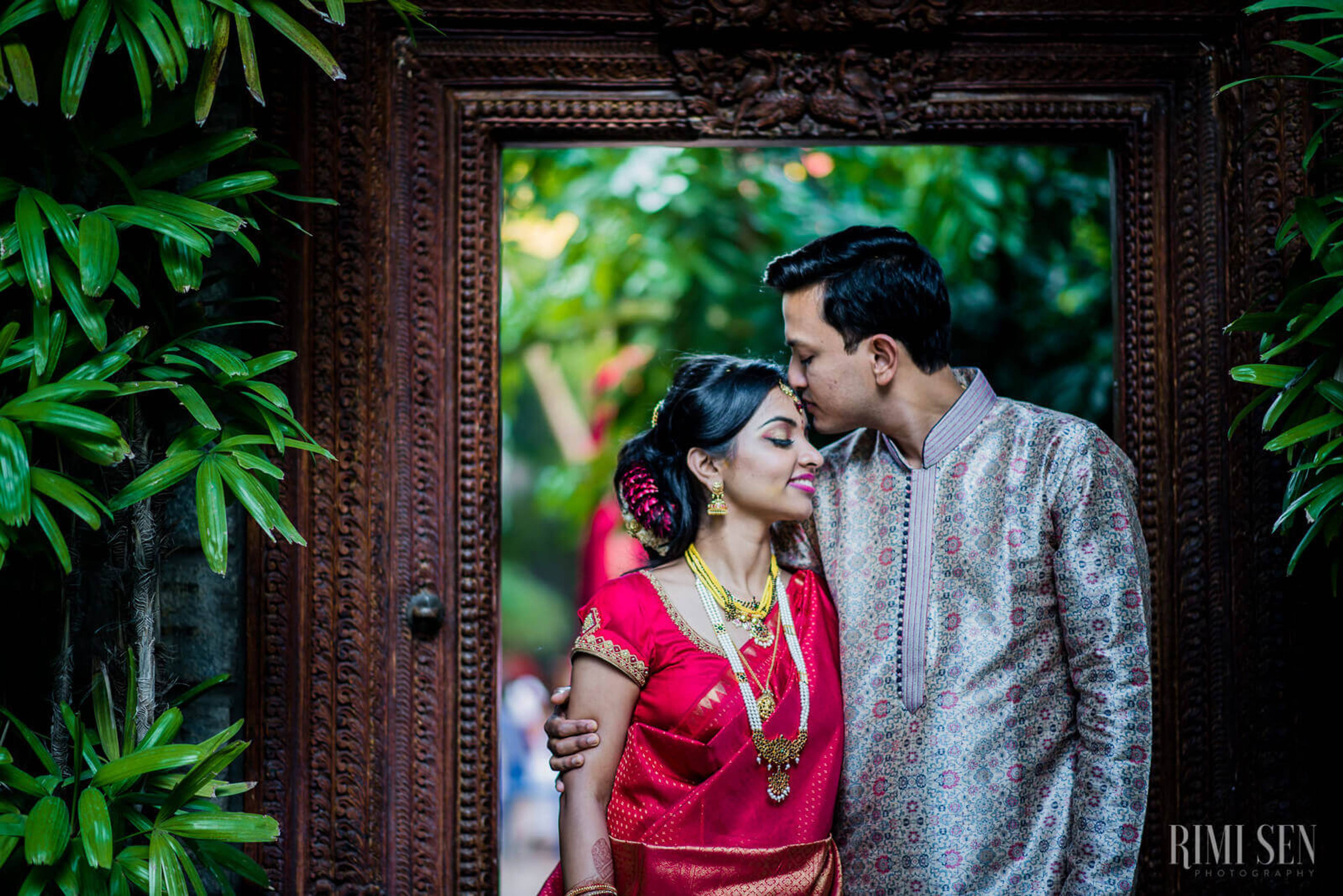 Pooja and Aniket embrace by an ornate wooden frame surrounded by lush green foliage in Bangalore