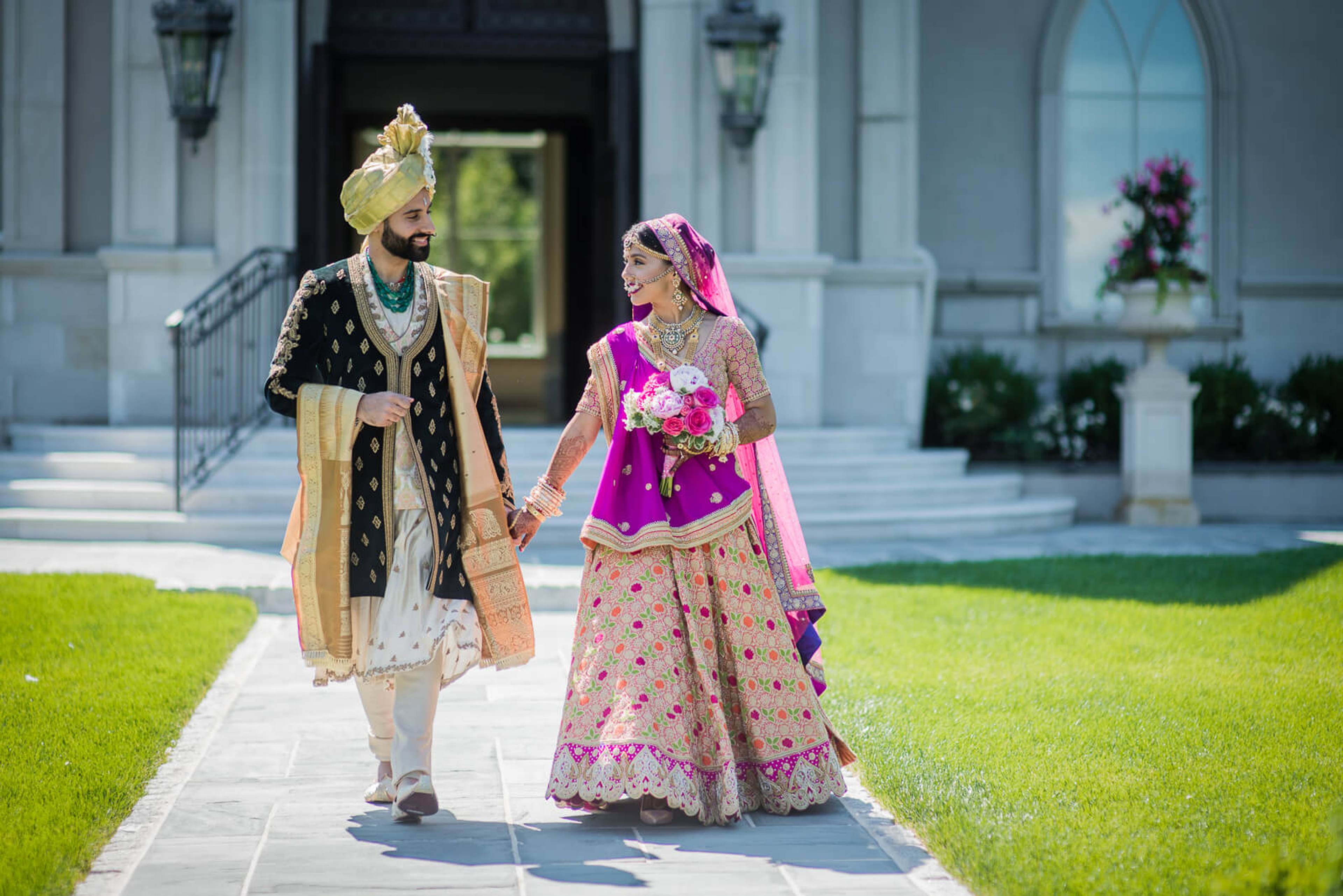 Barkha and Ankit walk hand in hand in traditional Indian attire on a sunlit path by Rimi Sen