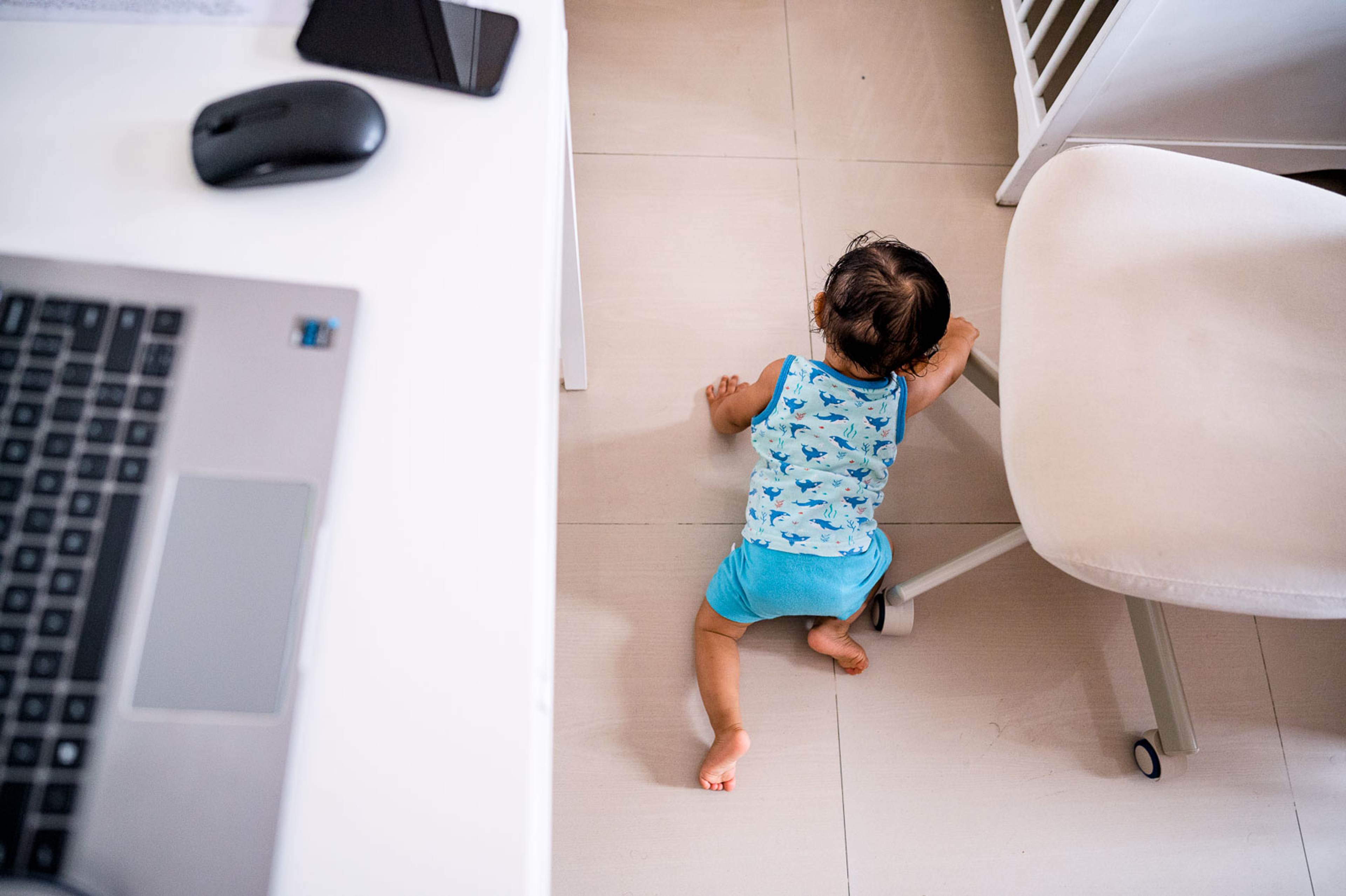 Toddler in blue print shirt crawling on tile floor near laptop and office chair, overhead view