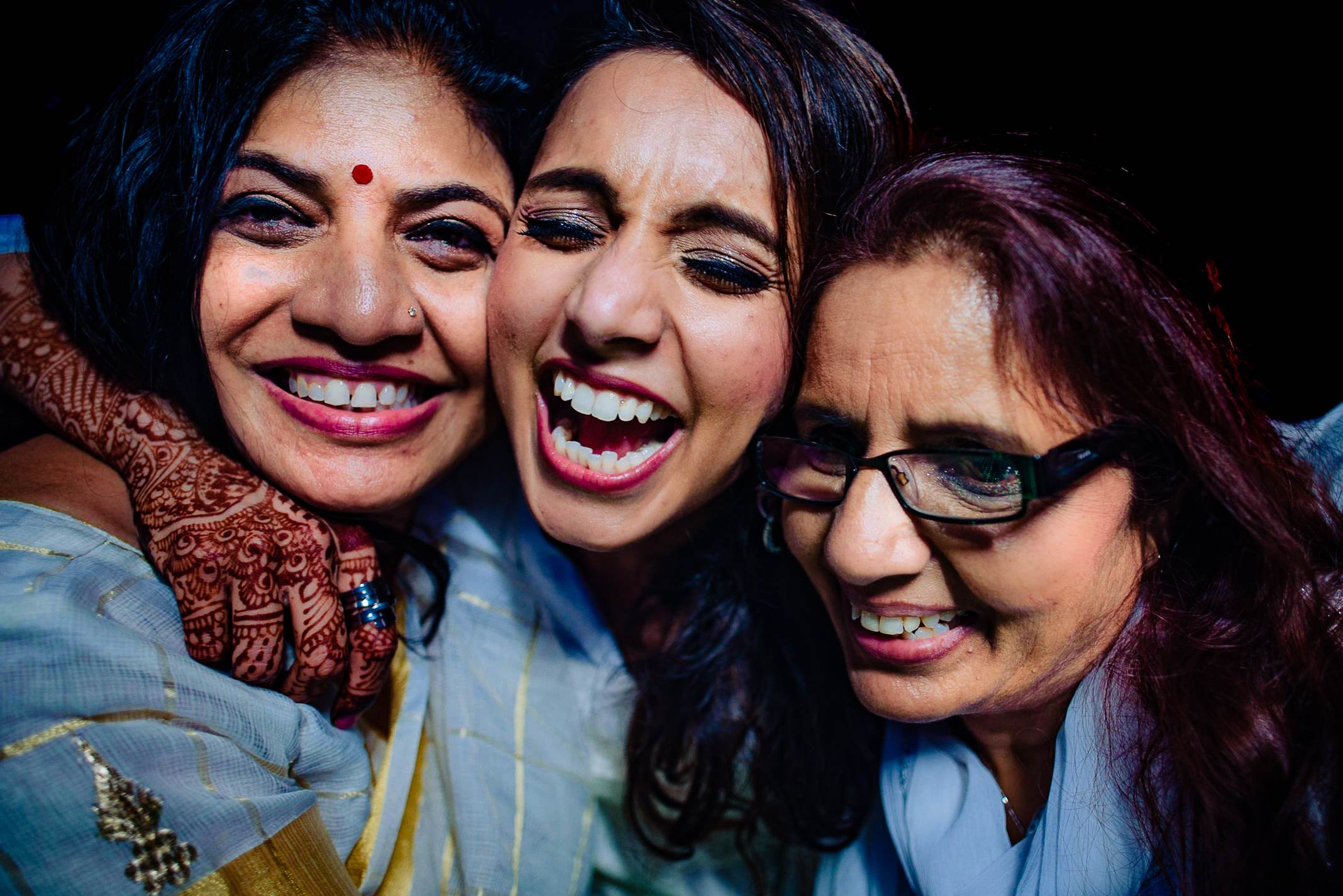 Three women laughing together in traditional Indian attire against dark background, Goa