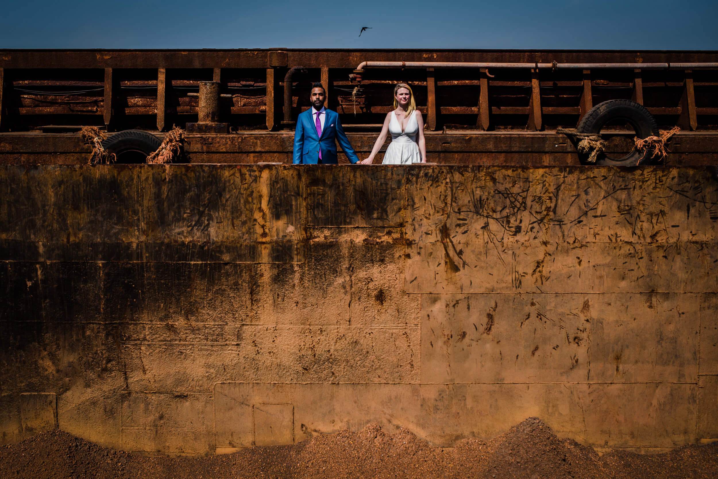 Sarah and Ravin hold hands on a weathered ship hull in Goa, clear sky behind them