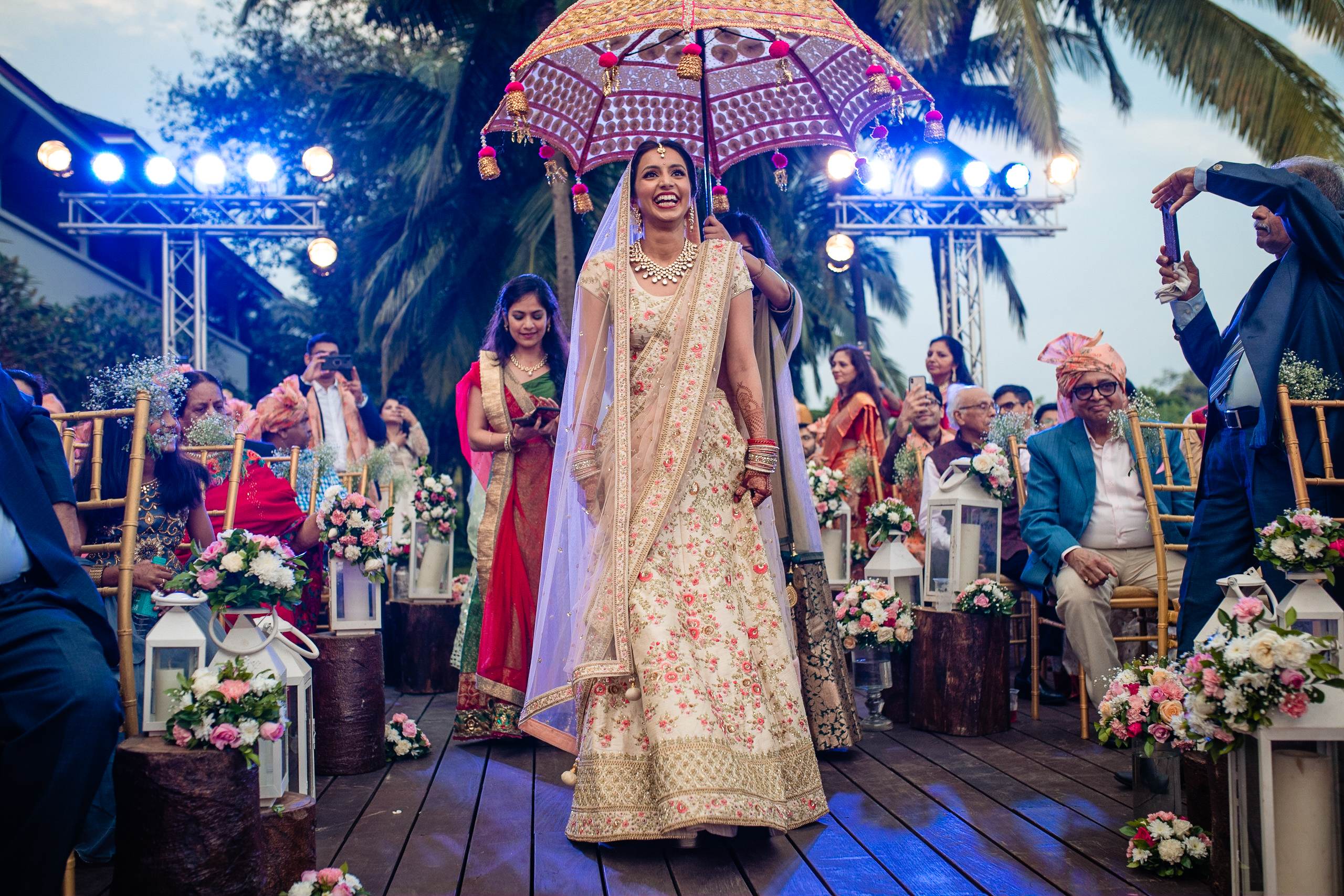 Avani walks under a pink patterned umbrella in ornate gold lehenga during her Goa wedding ceremony under evening lights
