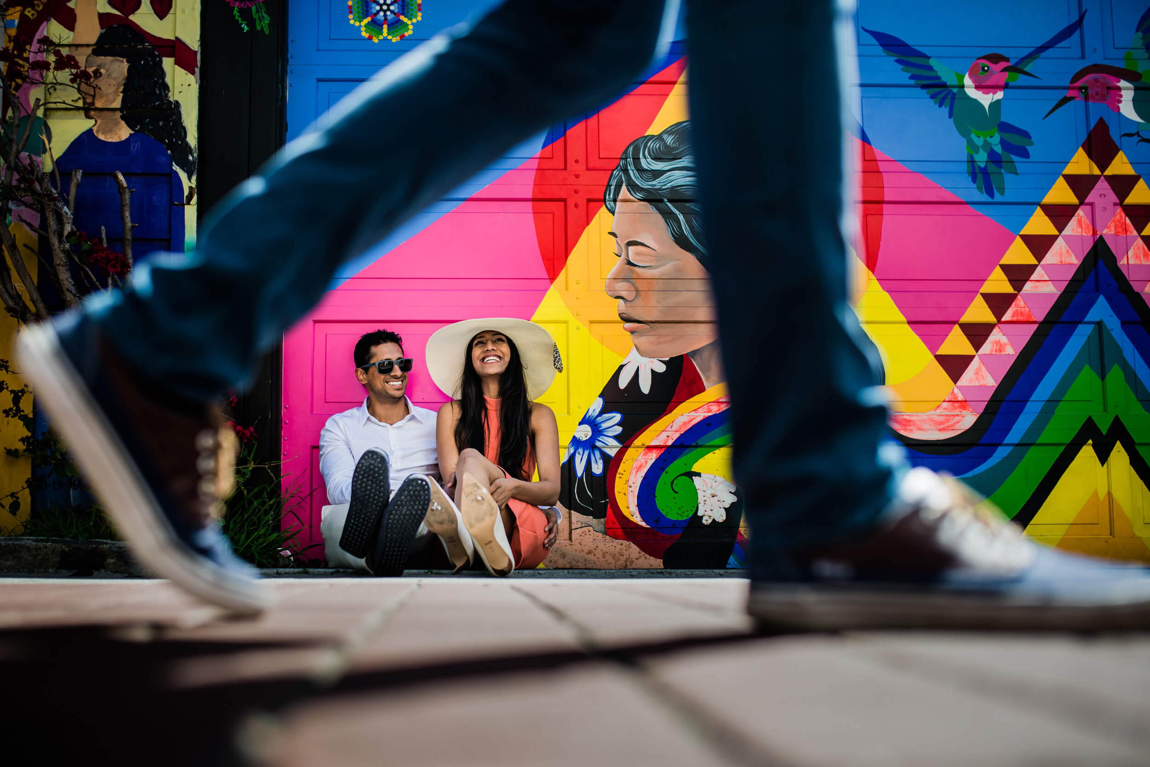 Anu and Raghu sit together in front of vibrant California street art mural, framed through chain-link fence