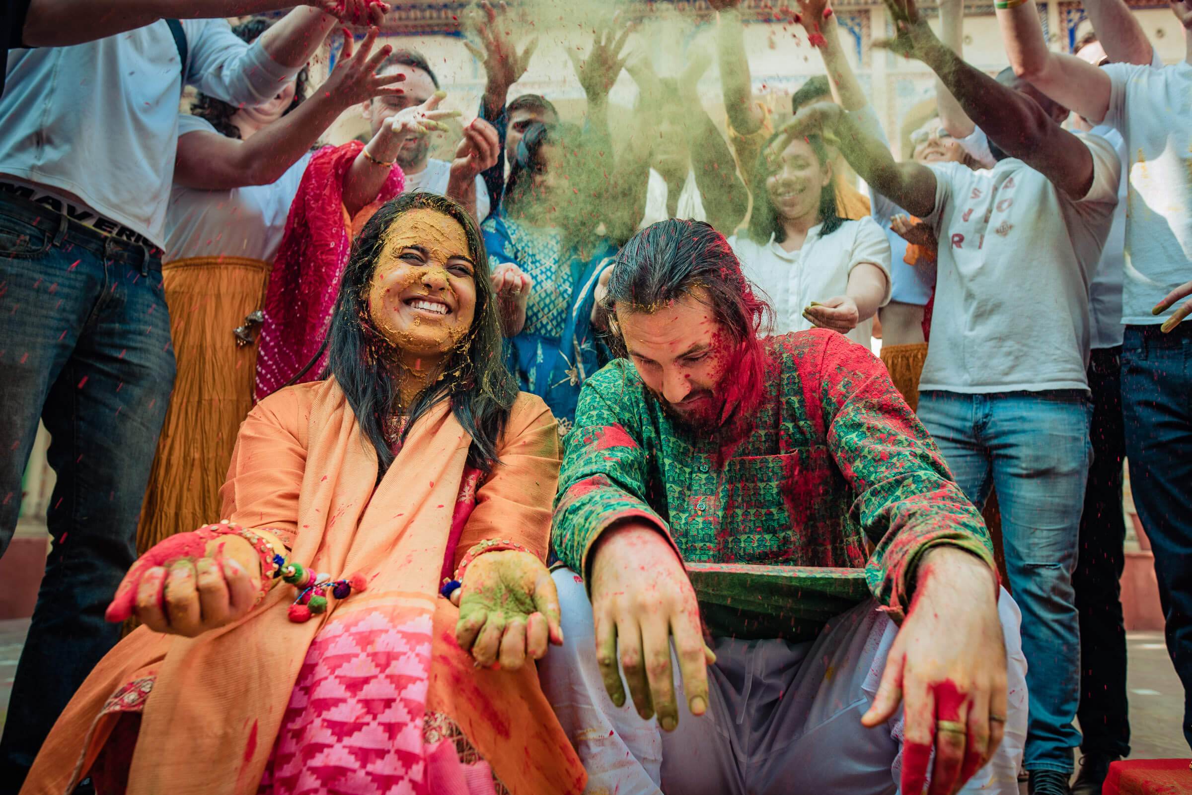 Maitrayee and Eric celebrate Holi in Rajasthan, covered in colored powder, surrounded by joyful guests