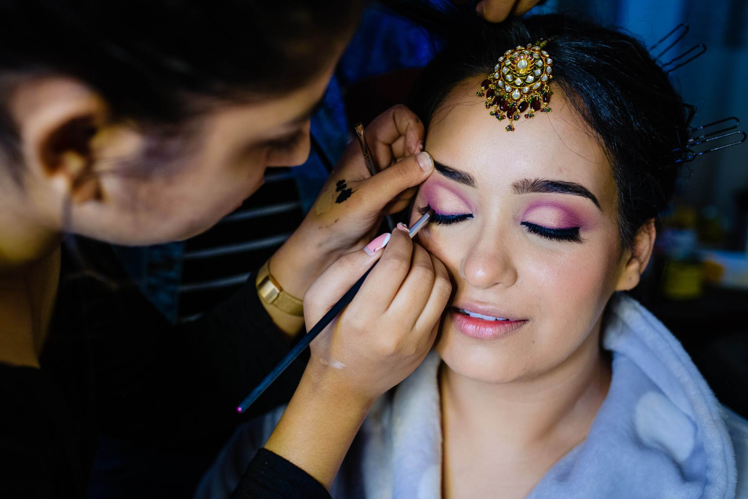 Makeup artist applies pink eyeshadow to bride with gold bindi in preparation for Rajasthan wedding