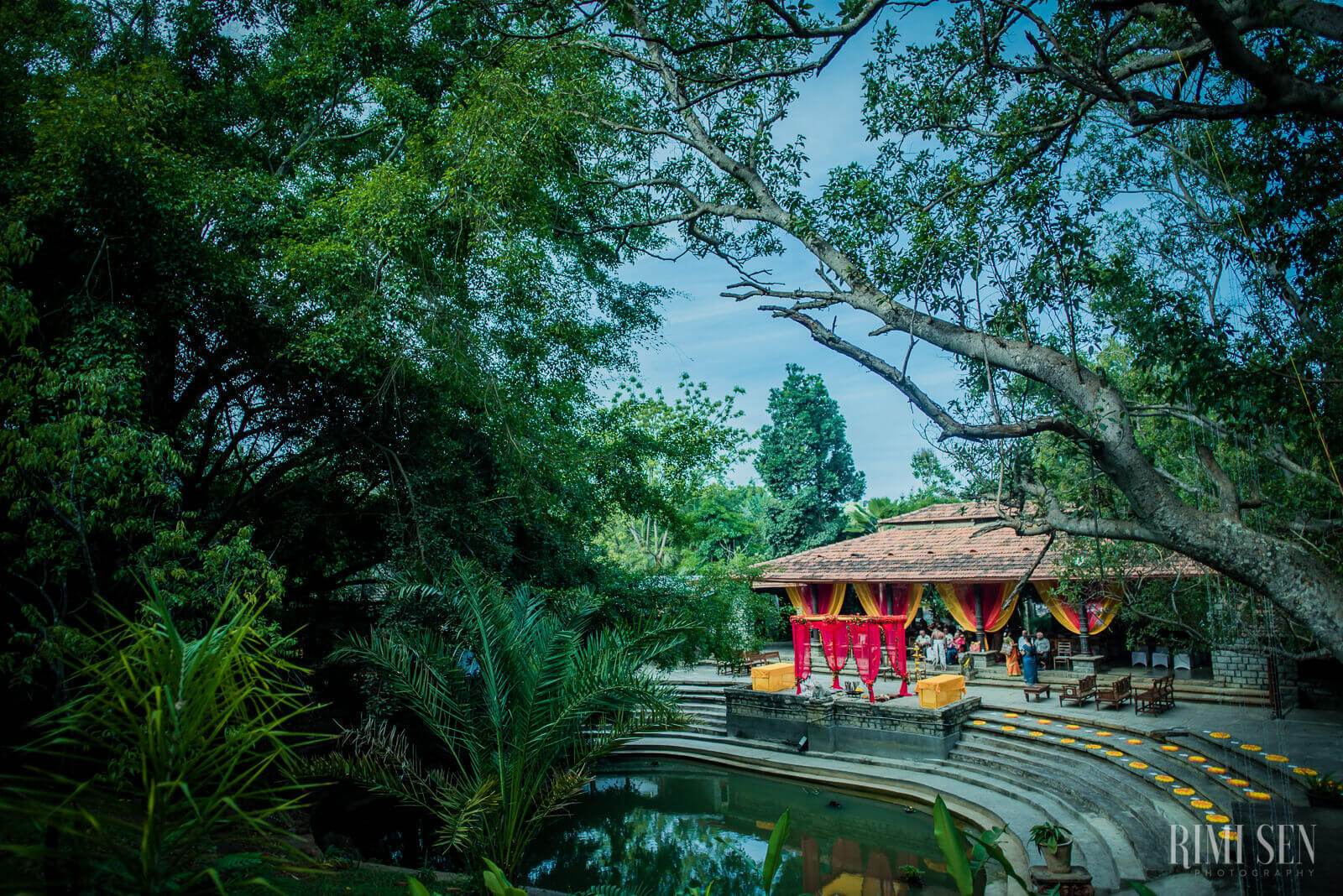 Red pavilion temple beside still water under lush green canopy trees, Bangalore