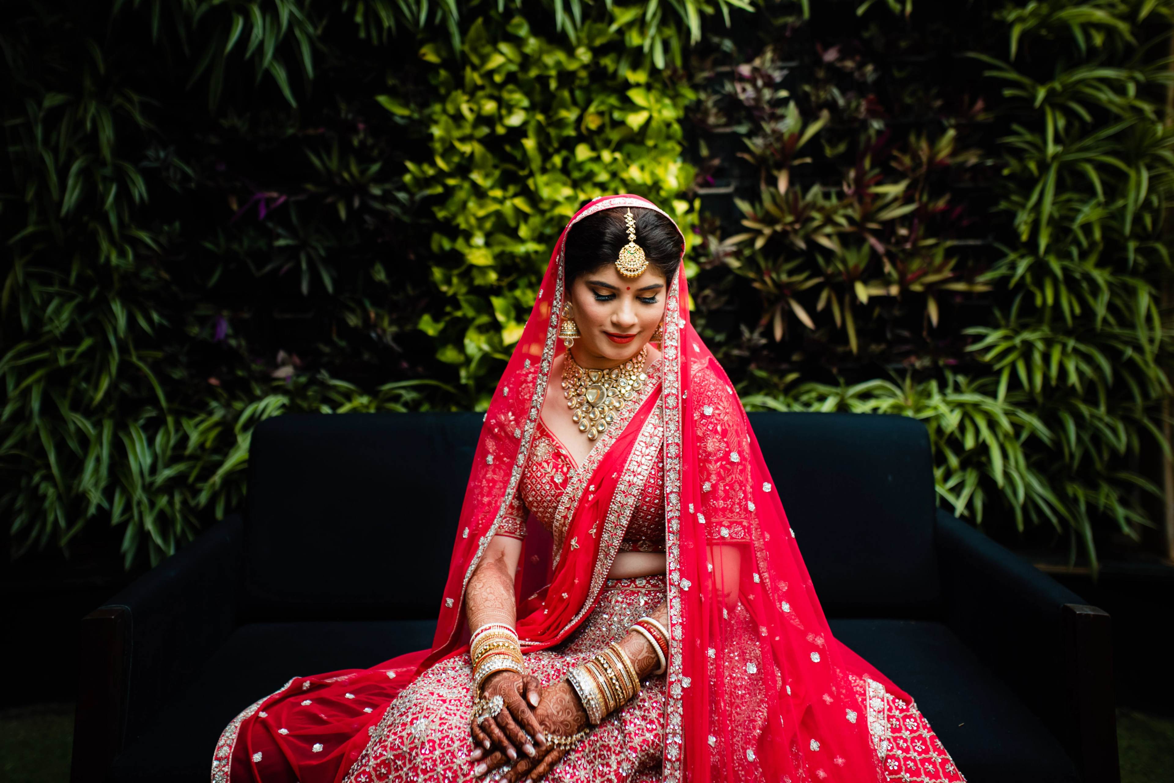 Woman in red bridal lehenga and veil seated against lush green foliage backdrop