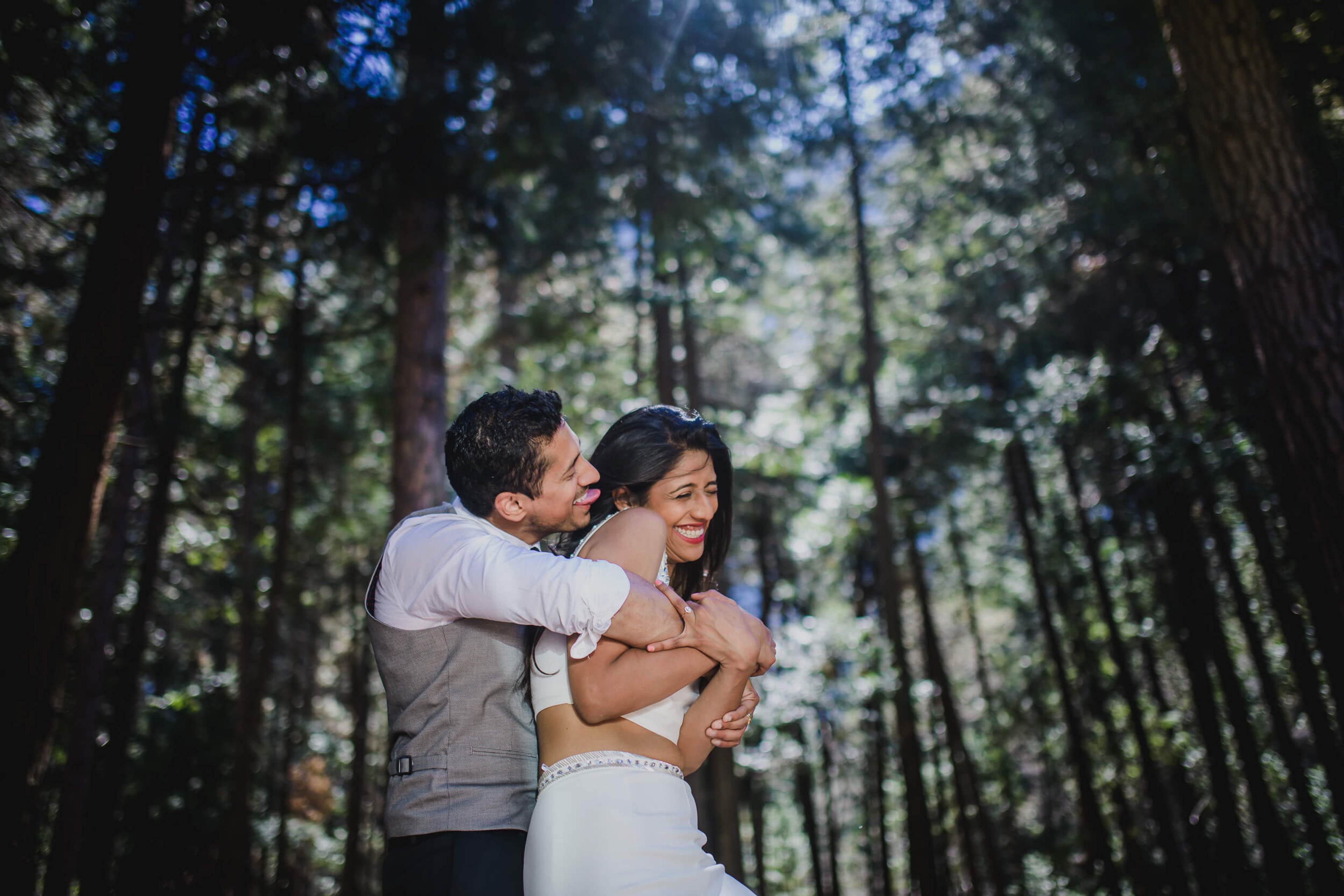 Anu and Raghu embrace in a sunlit forest path surrounded by tall trees in California