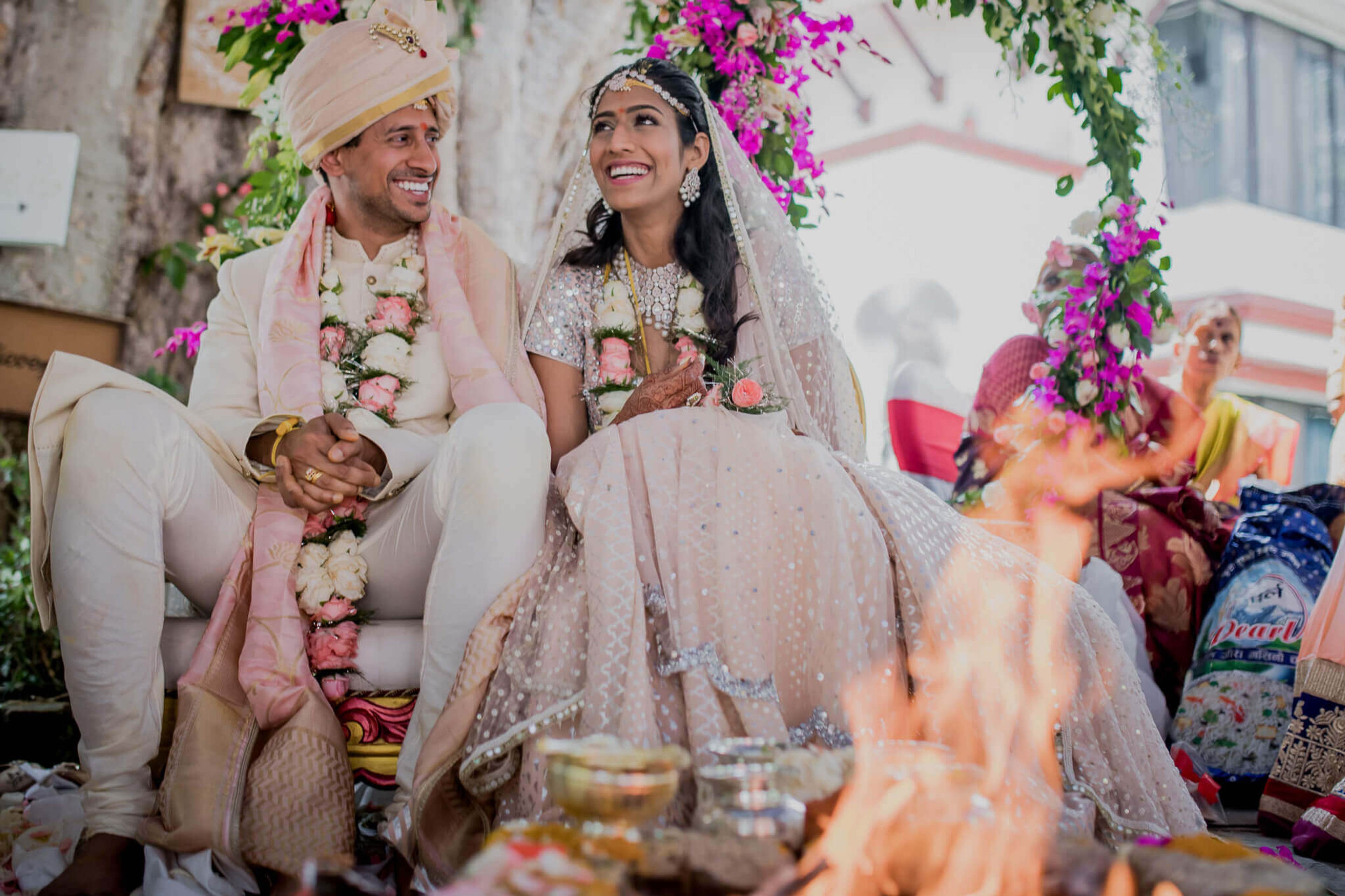 Anu and Raghu sit together in traditional wedding attire beneath magenta flowers, holding hands during their Nepal ceremony