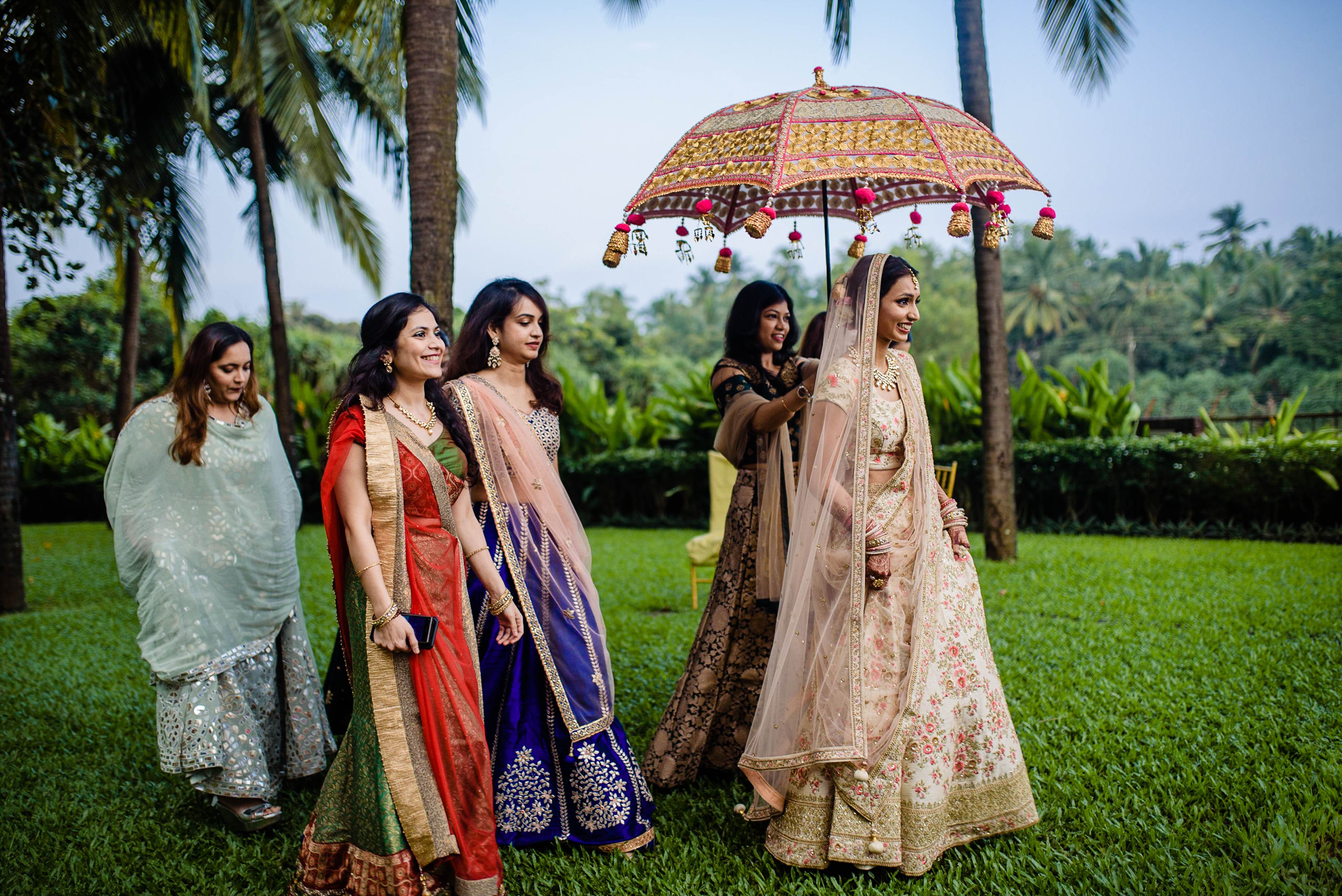 Avani and Srikant's wedding party in traditional attire with ornate umbrella on manicured lawn, Goa