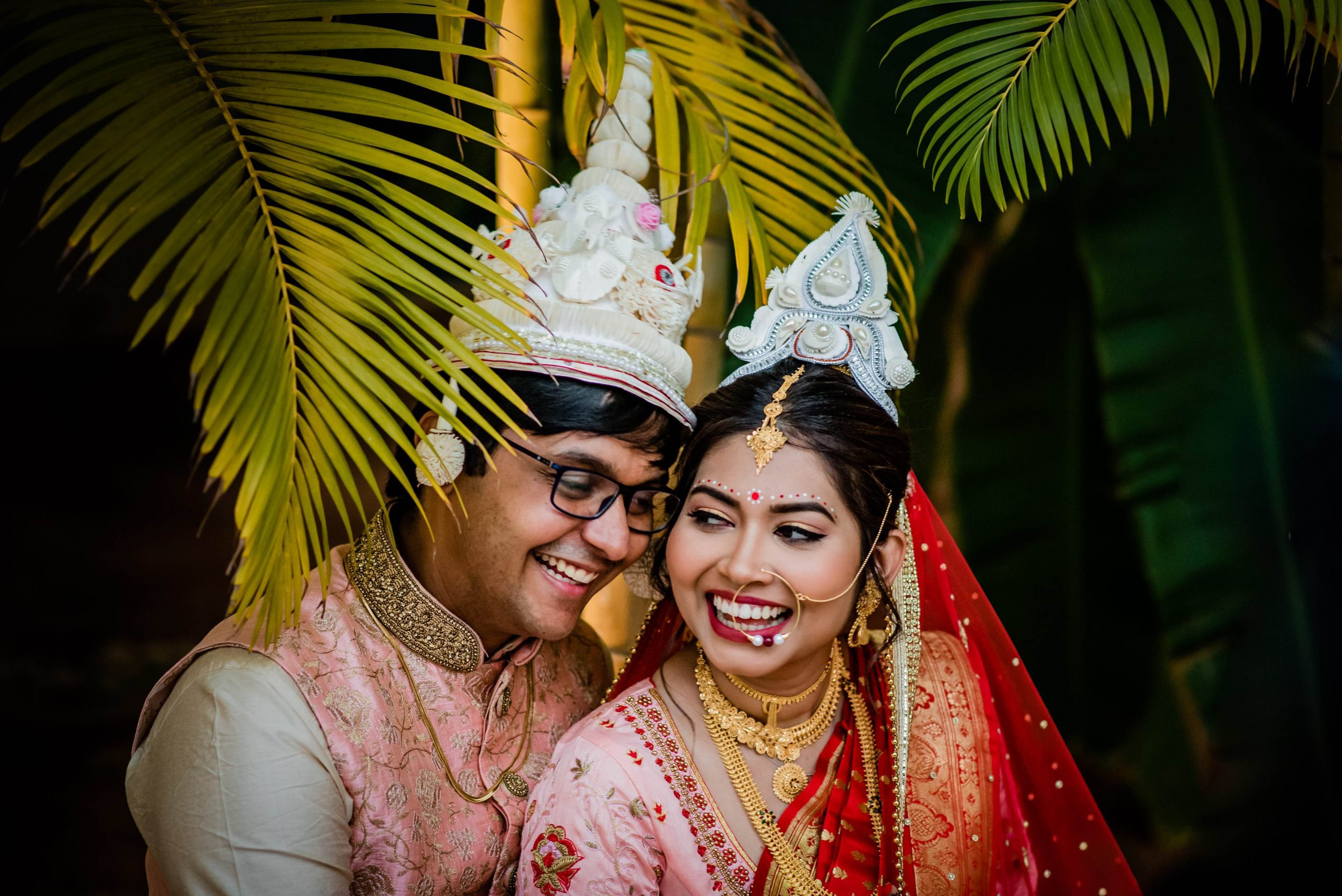 Couple in traditional attire and ornate headpieces smiling under palm fronds at night
