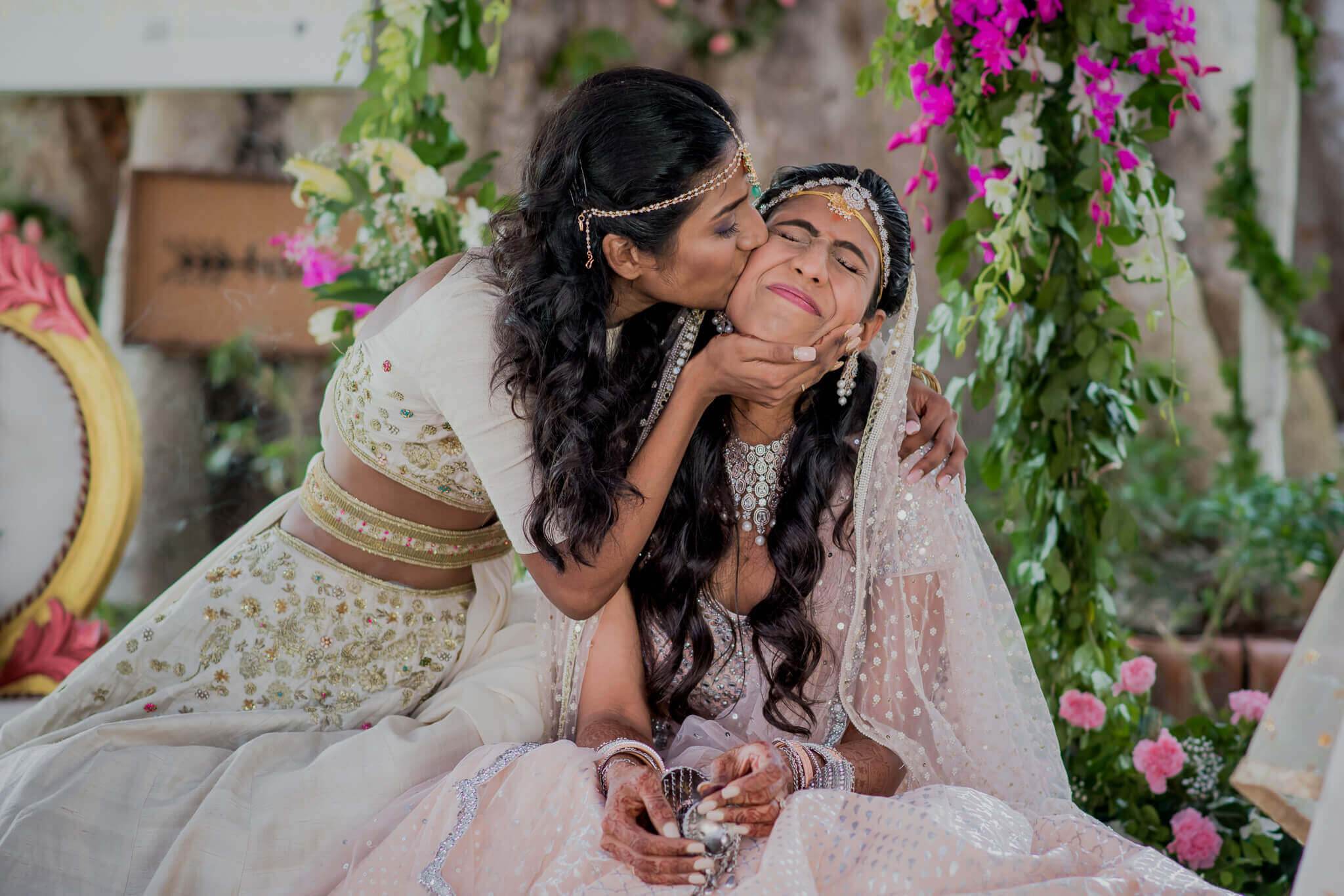 Anu kissing Raghu's cheek while seated among vibrant pink and white flowers in Nepal