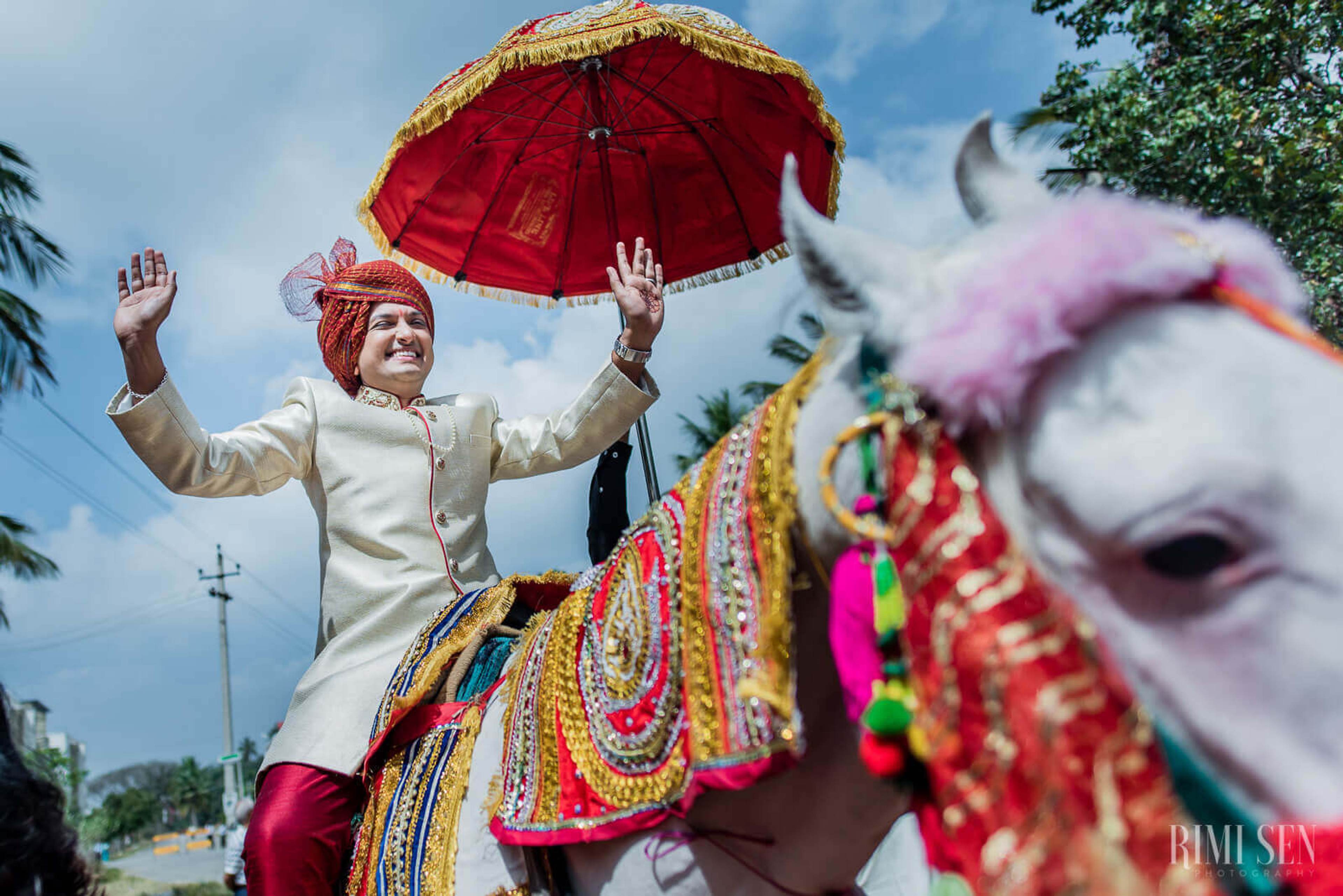 Groom in cream sherwani with red umbrella on decorated horse, Bangalore wedding celebration