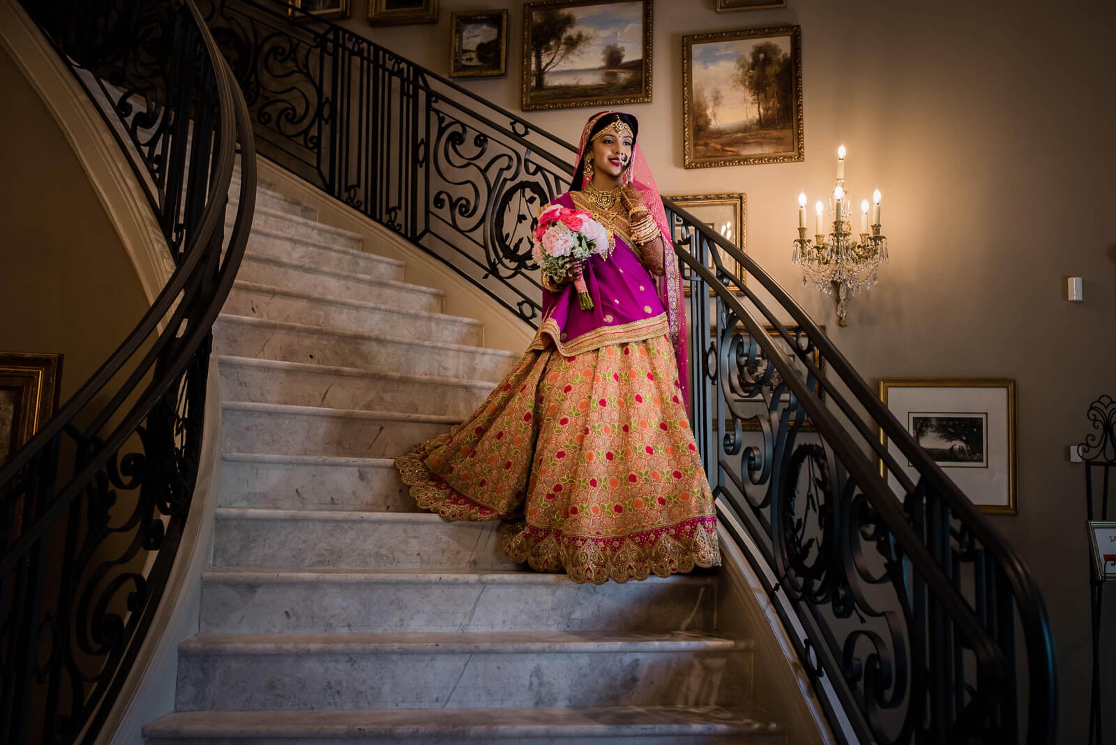 Barkha descends elegant staircase in vibrant pink and gold lehenga, holding floral bouquet in New York venue
